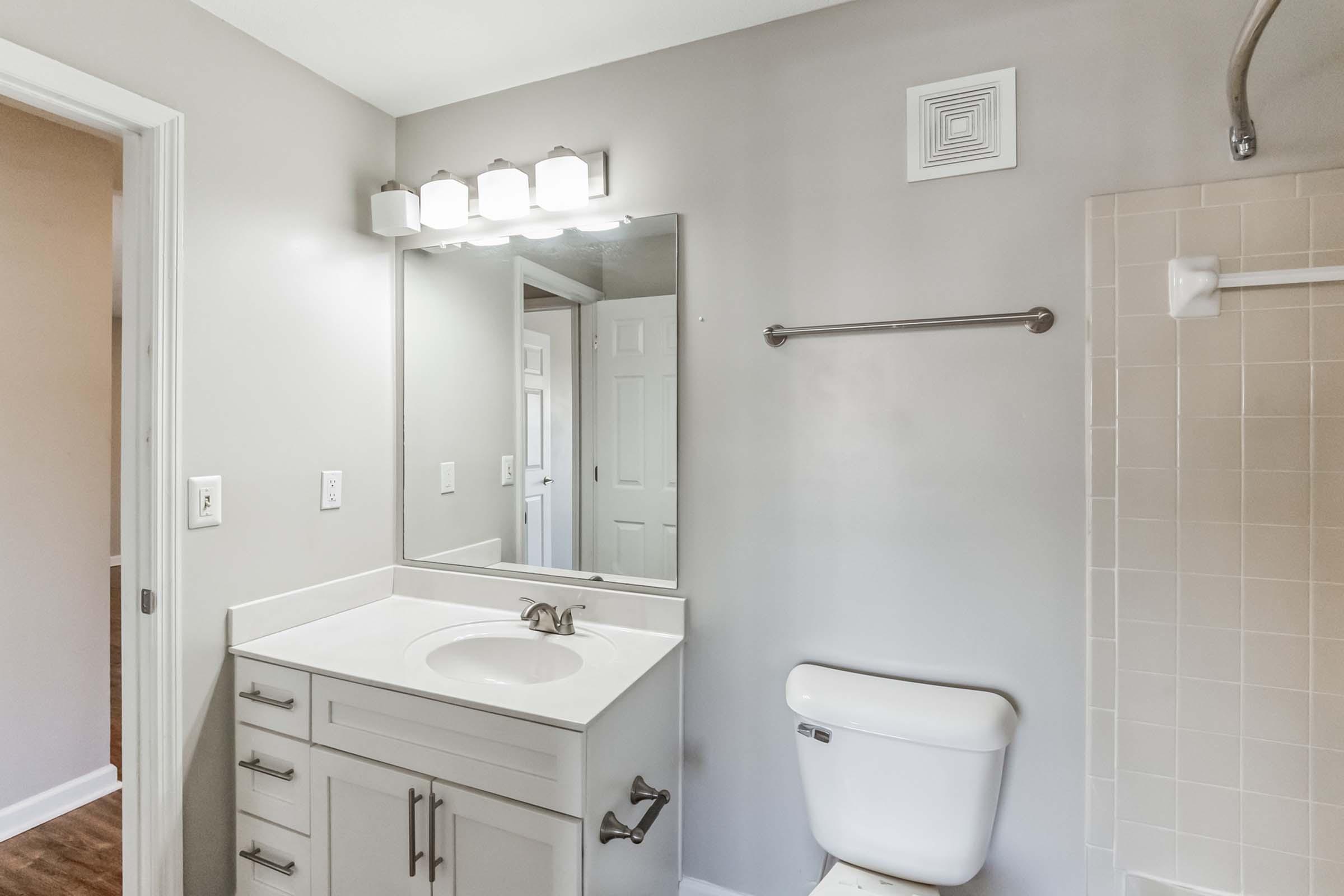 Modern bathroom with a light gray color scheme. It features a white sink with a stylish faucet, a large mirror above the sink, a towel bar, and a toilet. The shower area is visible, with a simple white wall. A door leads to another room, adding to the spacious feel. Bright lighting enhances the clean appearance.