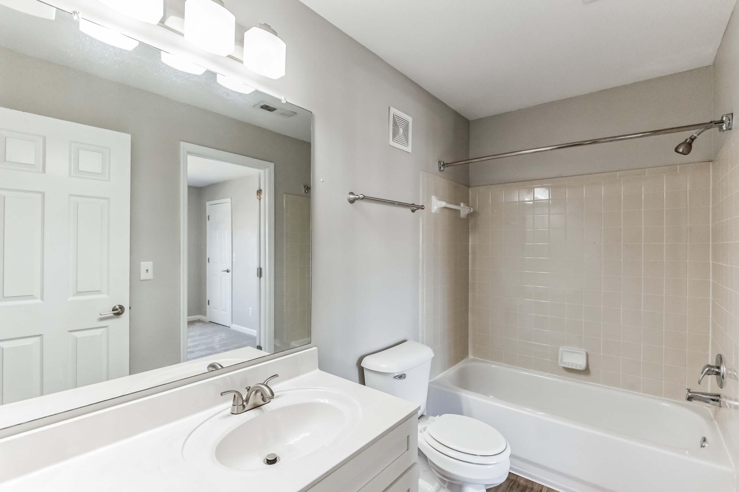 A modern bathroom featuring a white sink and countertop, a bathtub with a shower rod, and light beige tile walls. A large mirror reflects the space, which has neutral gray walls and a door leading to another room. Bright lighting from overhead fixtures enhances the clean and spacious feel of the area.