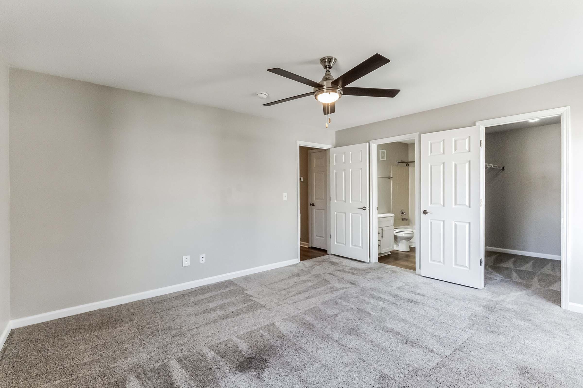 A spacious room featuring light gray walls and plush carpet, with a ceiling fan. Two doors lead to a bathroom and closet area, showcasing a bright and airy atmosphere ideal for relaxation or use as a bedroom. Natural light enhances the inviting ambiance.