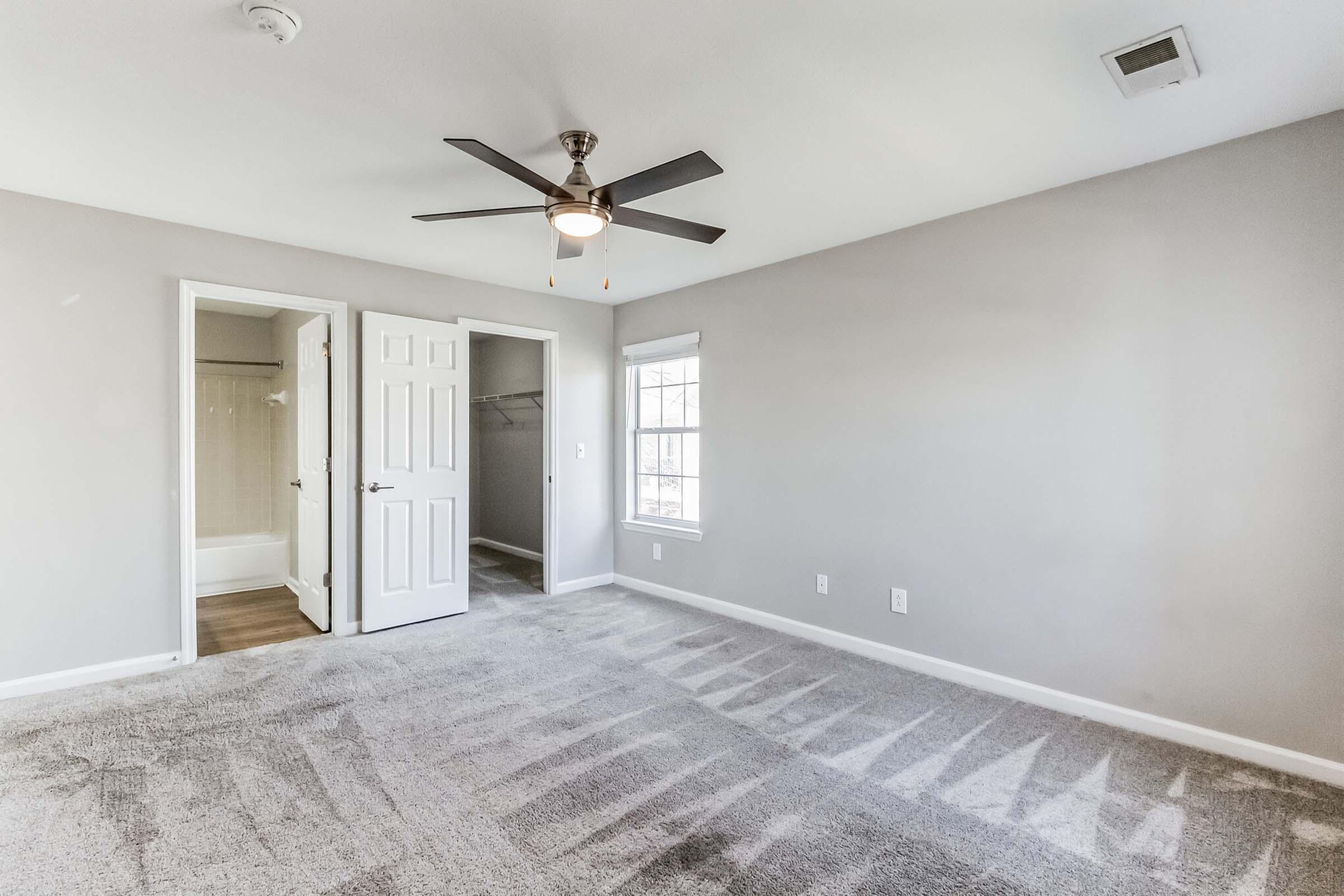 A spacious bedroom with light gray walls and carpet. The room features a ceiling fan, a window allowing natural light, and a closet with double doors. A bathroom entrance is visible on the left, and the overall atmosphere is bright and inviting.