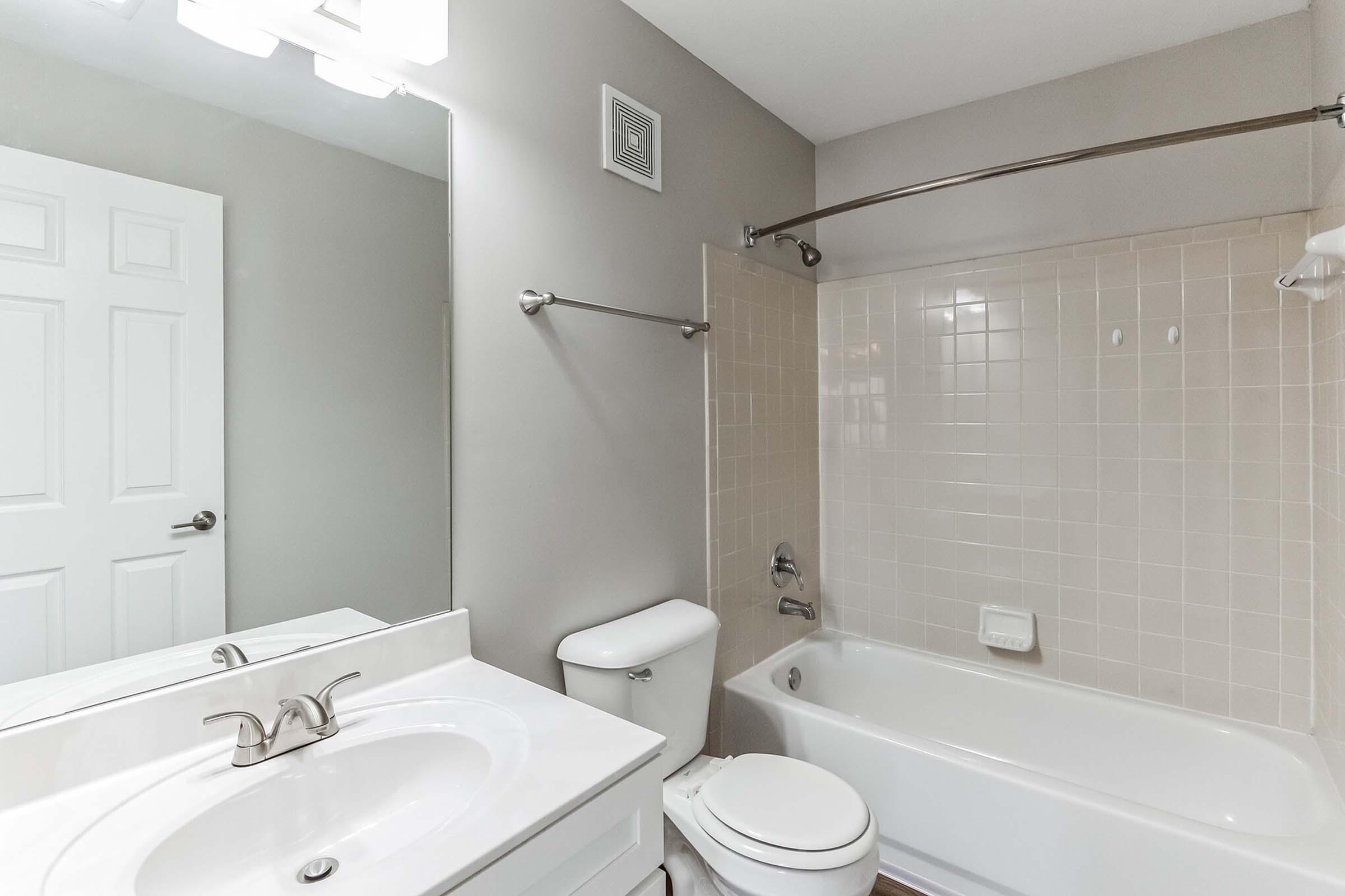 A clean, modern bathroom featuring a white sink with a mirror above, a toilet, and a bathtub with a shower curtain. The walls are painted a neutral gray, and the tile in the bathtub area is light-colored. A door is visible on the left side of the image. Soft lighting enhances the bright atmosphere.