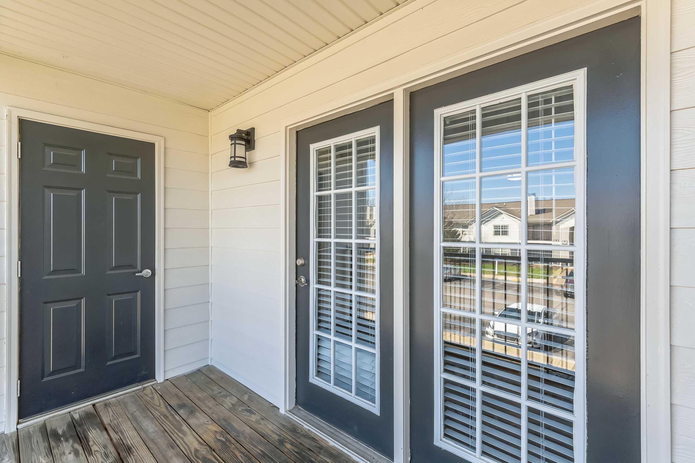 Front porch view featuring a wooden floor and two glass-paneled doors with dark frames. There is a dark door on the left and a light fixture above. The background shows a residential area with houses visible through the windows.