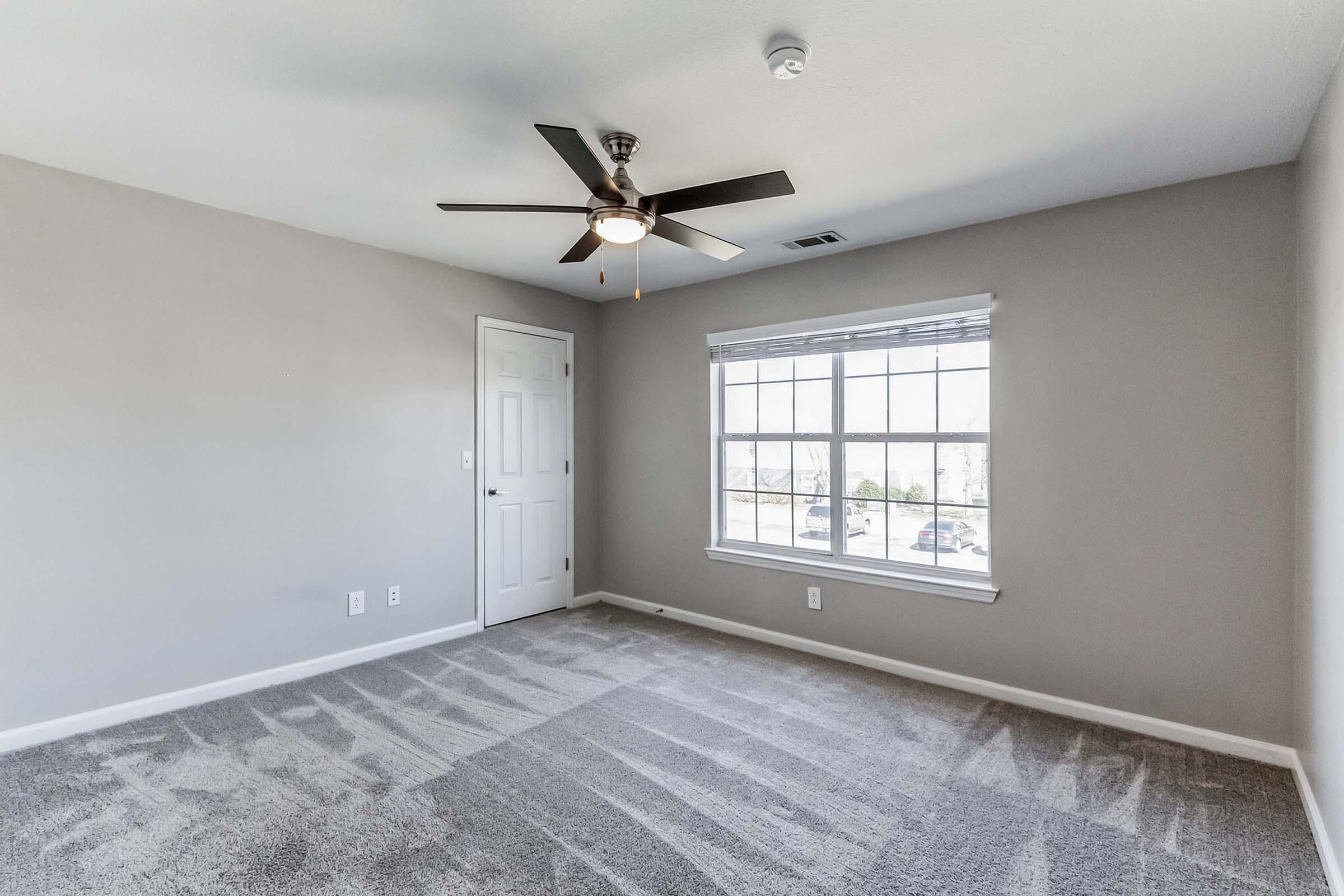 A clean, empty bedroom with light gray walls and a ceiling fan. The room features a large window allowing natural light, a white door, and freshly carpeted flooring with a light texture.