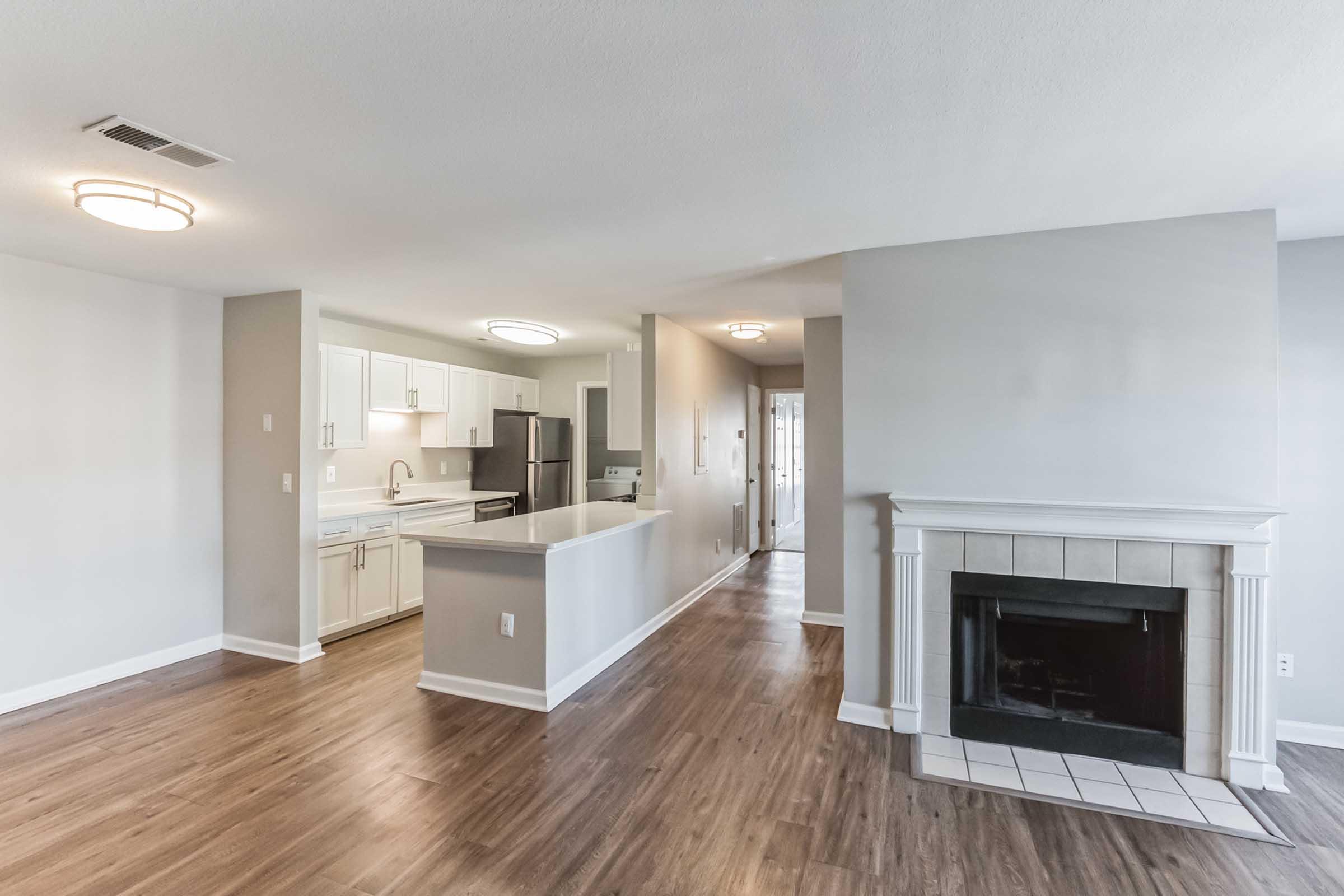 A spacious, modern living area with wooden floors, featuring a kitchen with white cabinets and stainless steel appliances on the left. There is a cozy fireplace in the living room area. Bright overhead lights illuminate the open space, creating a welcoming atmosphere.