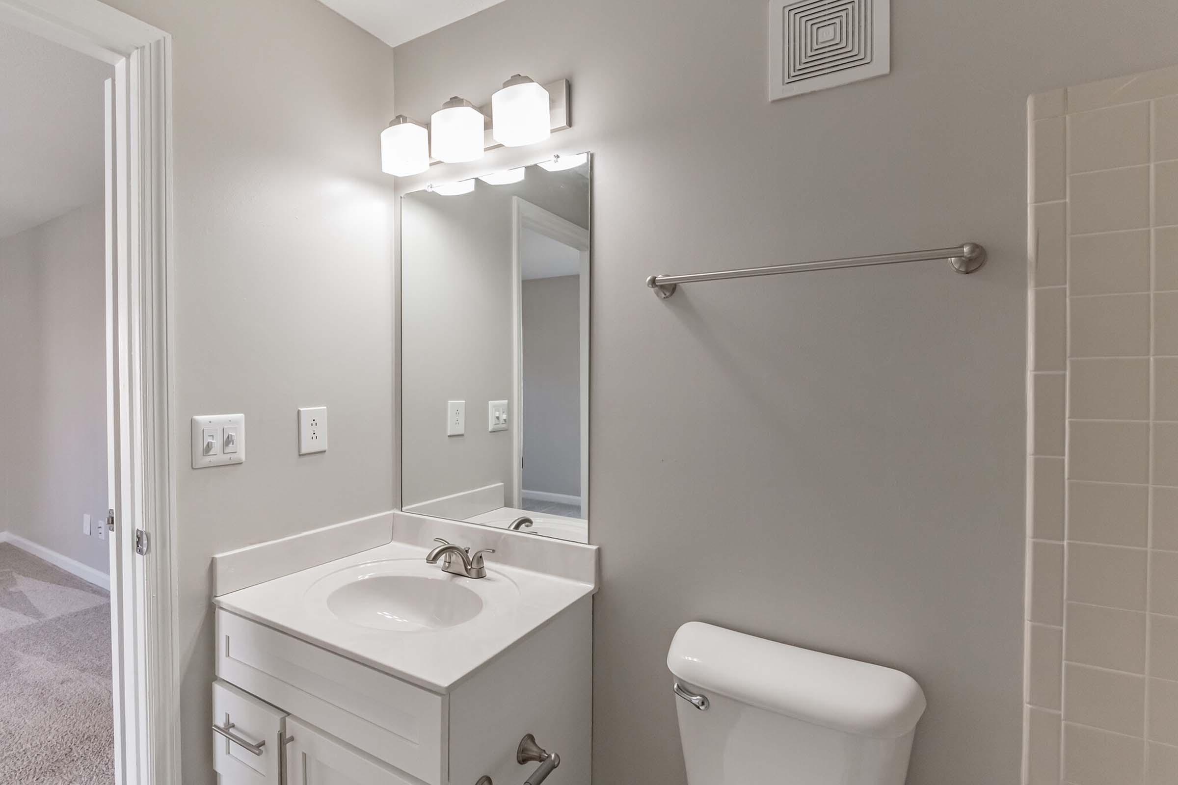 A modern bathroom featuring a white vanity with a sink, a large mirror above, and three light fixtures. A towel bar is mounted on the wall, and a toilet is placed nearby. The walls are painted a light gray, and the flooring is carpeted.
