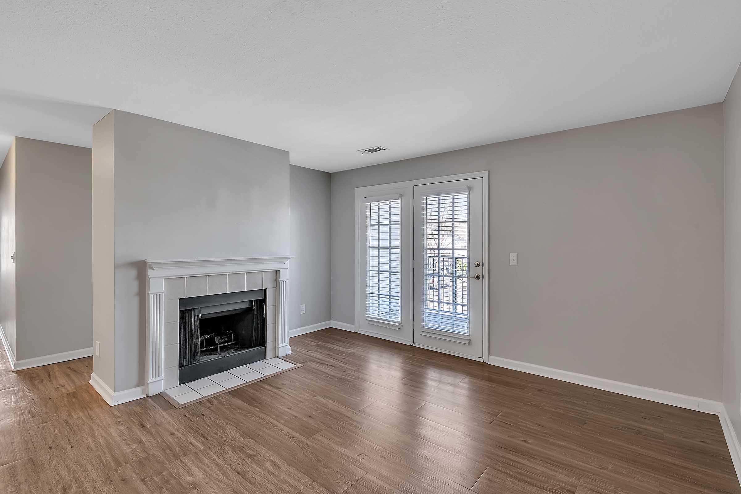 A spacious living room with light gray walls and hardwood flooring. The room features a white fireplace with a tiled base and large double doors leading to a balcony, adorned with white shutters. Natural light fills the space, enhancing the open and inviting atmosphere.