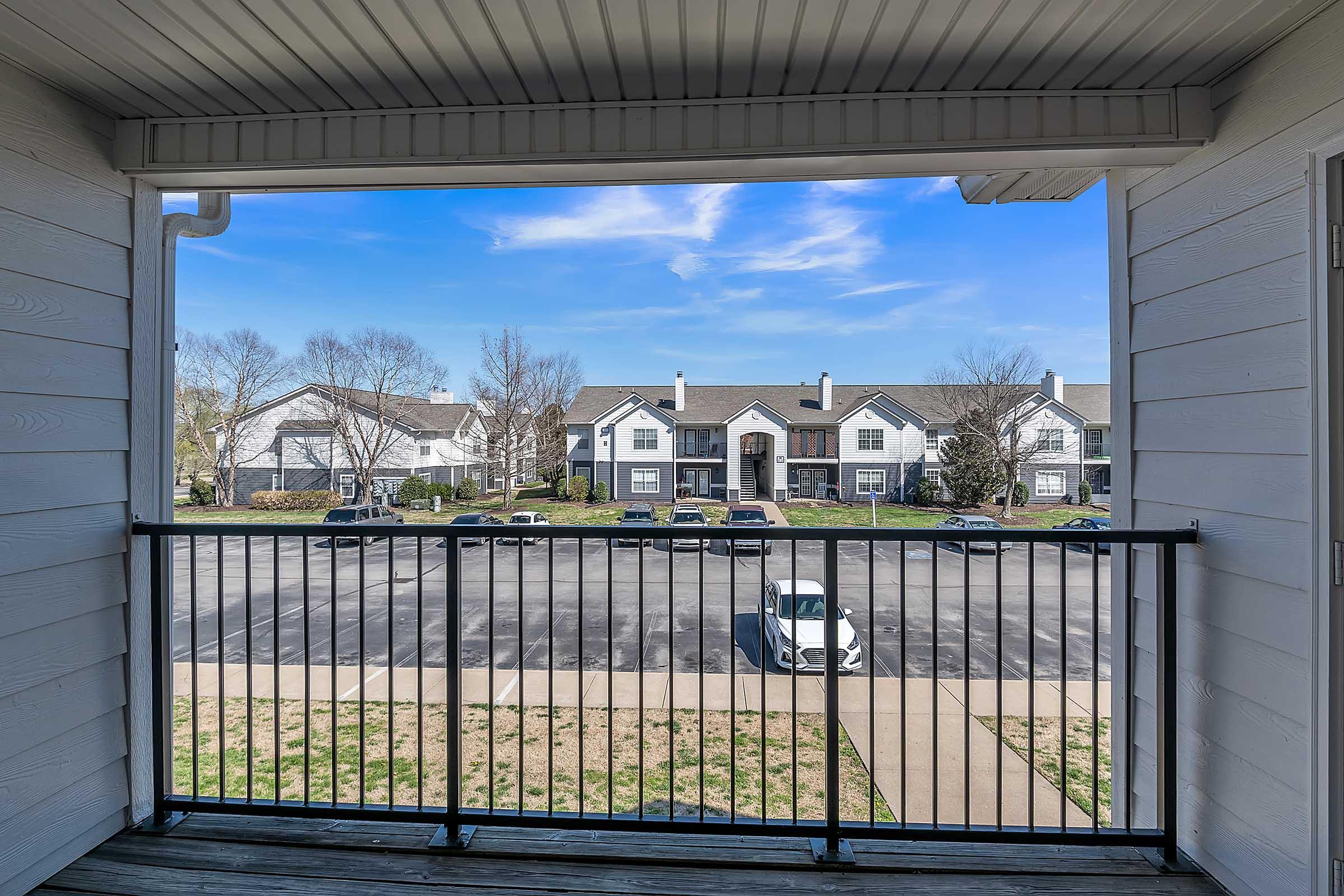 View from a balcony overlooking a parking lot and residential buildings. The sky is clear with a few clouds, and grass is visible in the foreground.