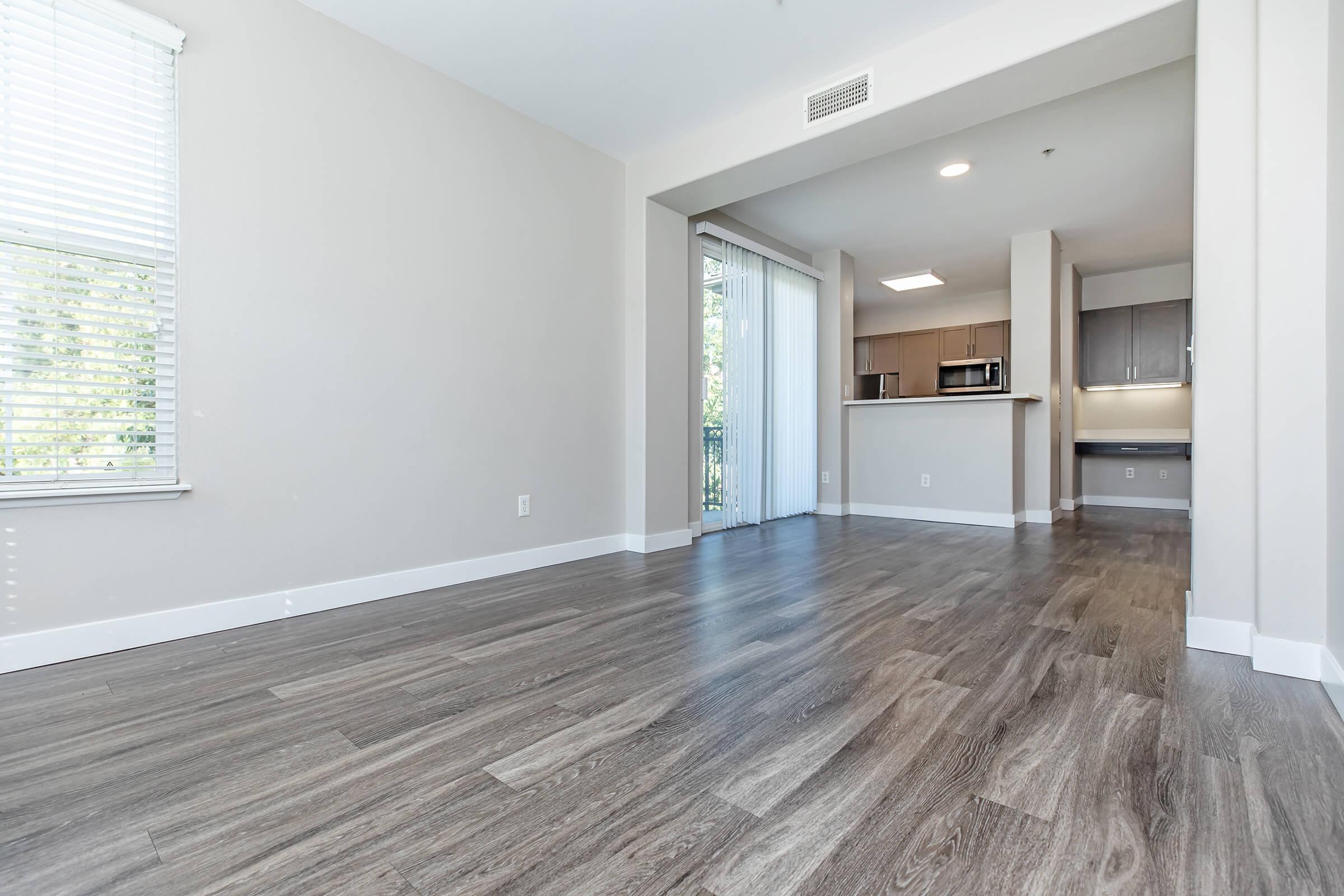 A spacious, well-lit living area with large windows, featuring modern wood flooring and neutral-colored walls. The adjacent kitchen is partially visible, showcasing contemporary cabinetry and appliances. Natural light filters in, creating a warm and inviting atmosphere.