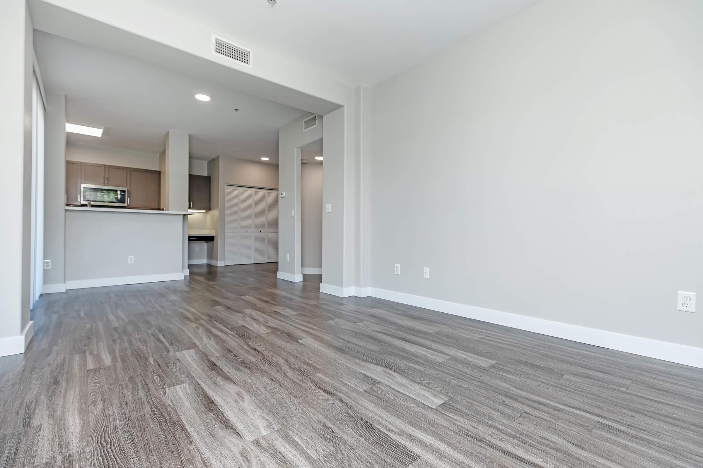 Empty interior space of a modern apartment featuring light gray walls, wood-like laminate flooring, and an open layout. In the background, a kitchen area with cabinets and counter space is visible, while a closet door is seen on the right. The overall atmosphere is bright and spacious, with plenty of natural light.