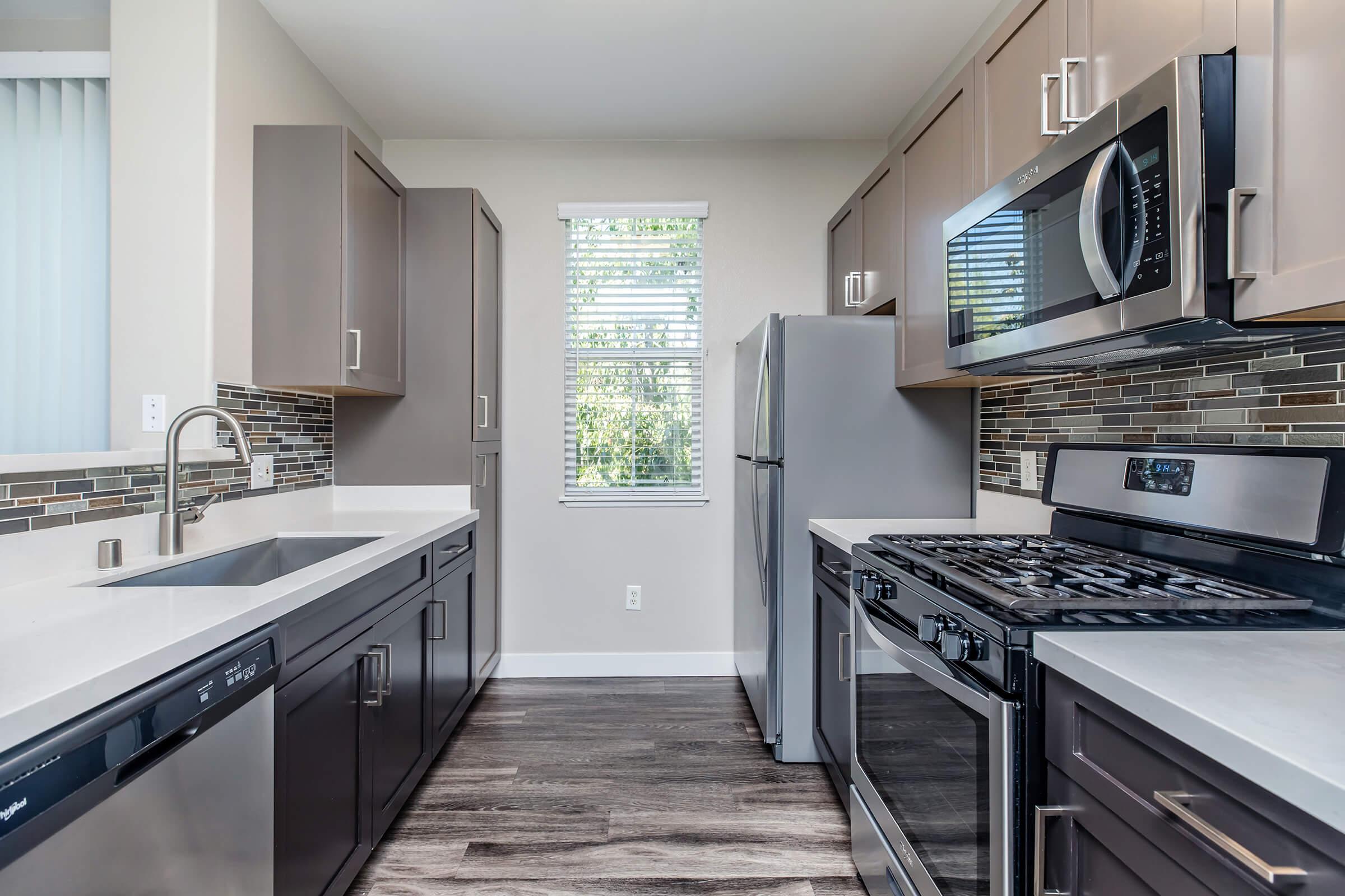 Bright modern kitchen featuring dark cabinetry, stainless steel appliances including a microwave and stove, and a spacious countertop. Natural light streams in through a window with blinds, and the floor is covered with wood-like tiles. A stylish backsplash adds a contemporary touch.