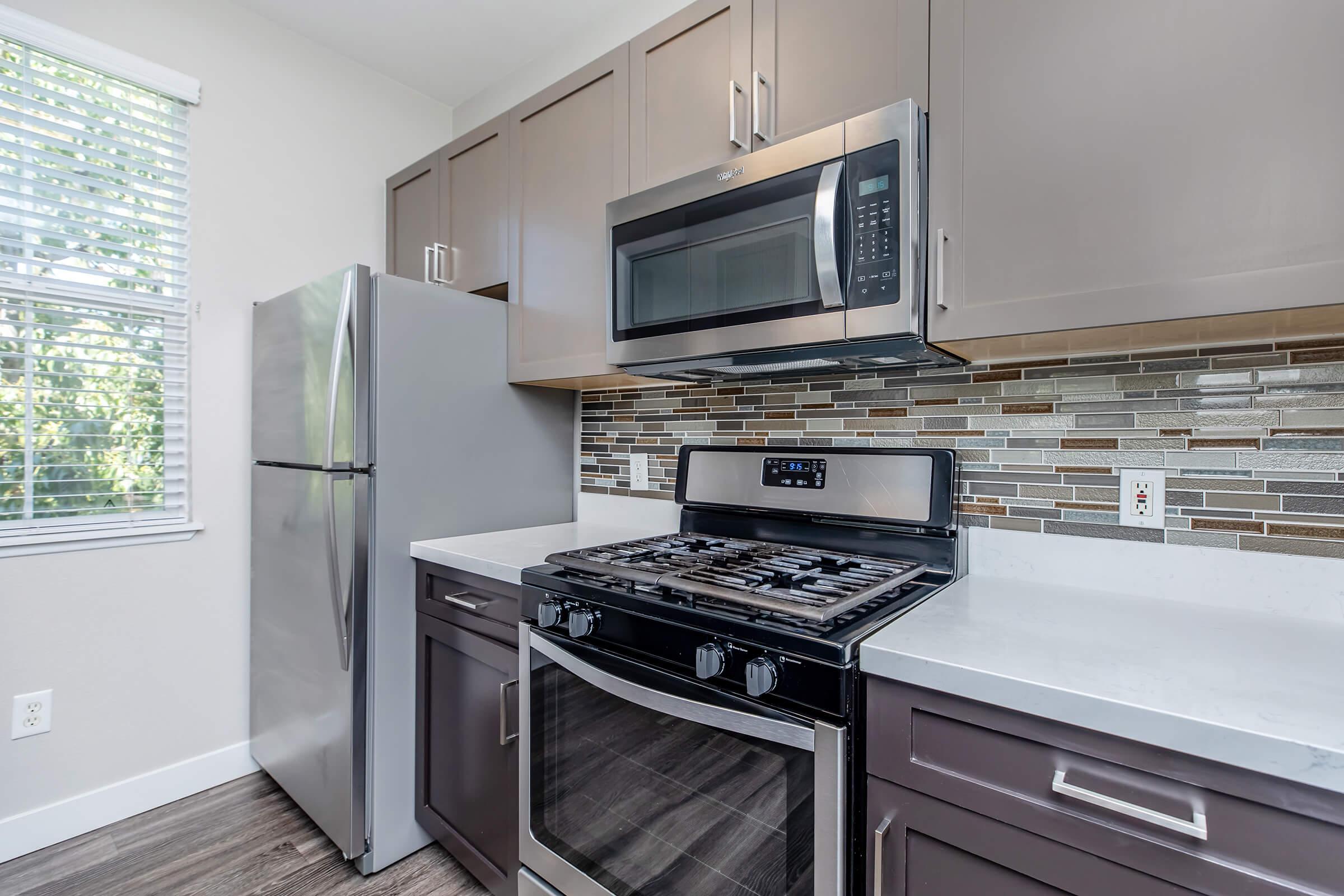 A modern kitchen featuring stainless steel appliances including a refrigerator, microwave, and gas stove. The cabinets are a mix of light and dark wood, with a stylish backsplash made of mosaic tiles. Natural light comes through a window with white blinds, illuminating the countertop and fixtures.