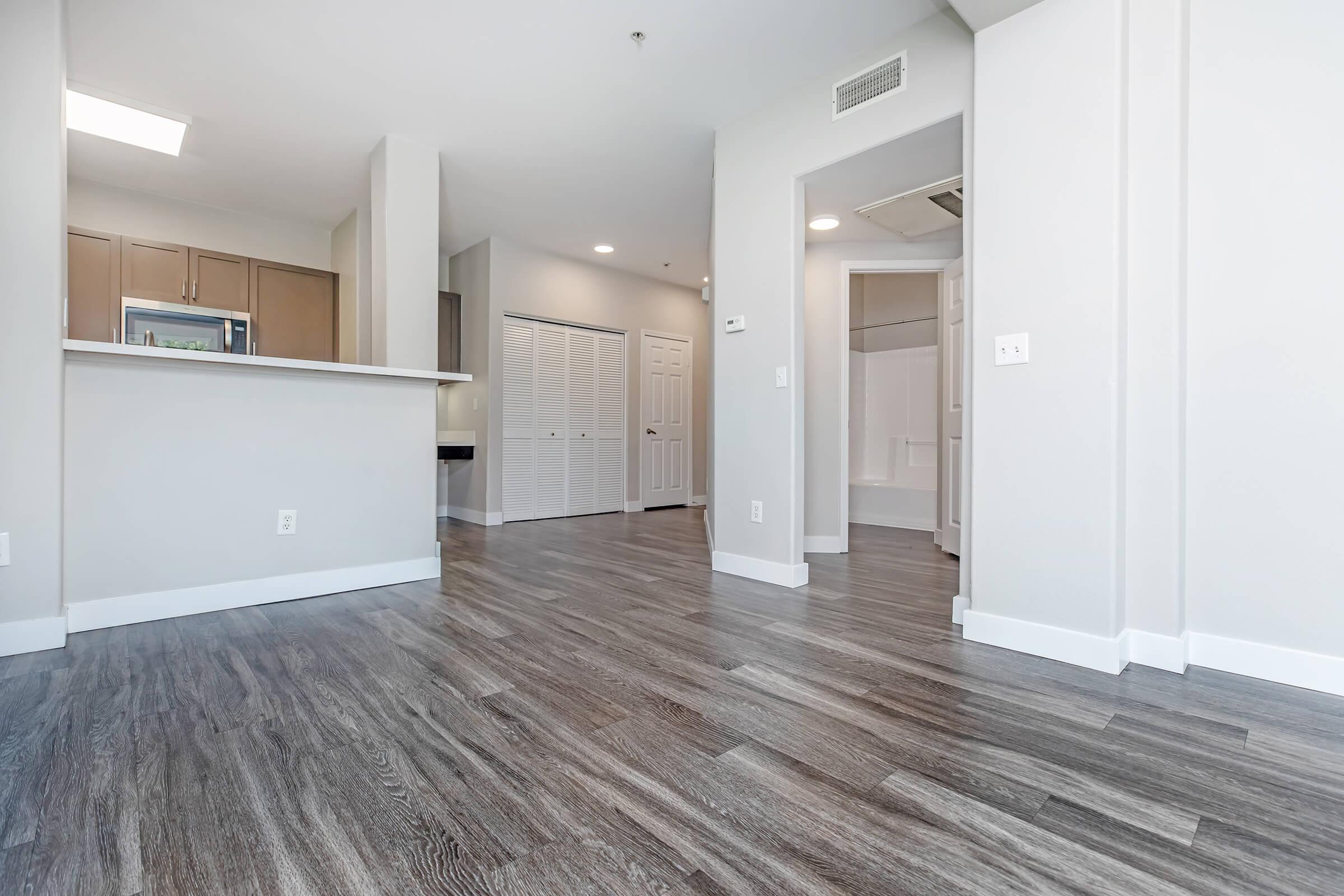 A spacious, modern interior of an apartment showcasing light-colored walls, sleek hardwood flooring, and a partially open kitchen area with a breakfast bar. The layout is bright and inviting, featuring a hallway leading to a bathroom and an adjacent closet space. Natural light illuminates the room from the ceiling.