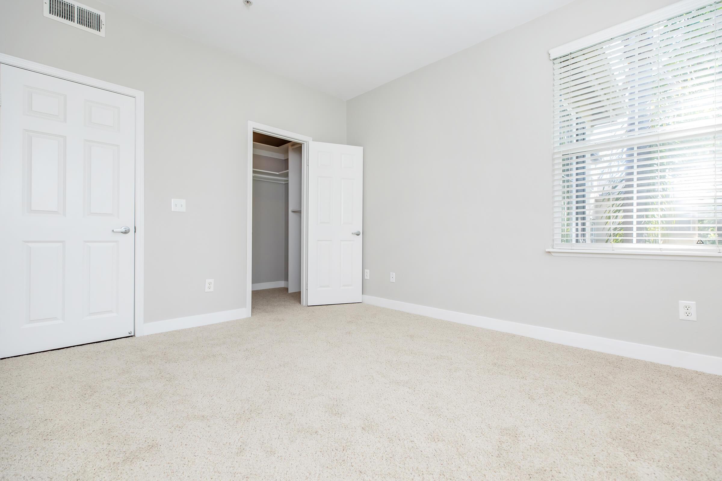 A well-lit, empty room with light gray walls and beige carpet. One wall has a window with blinds, letting in natural light. Opposite, there are two closed white doors: one leading to a closet and the other to a space outside the room, creating a clean and spacious feel.