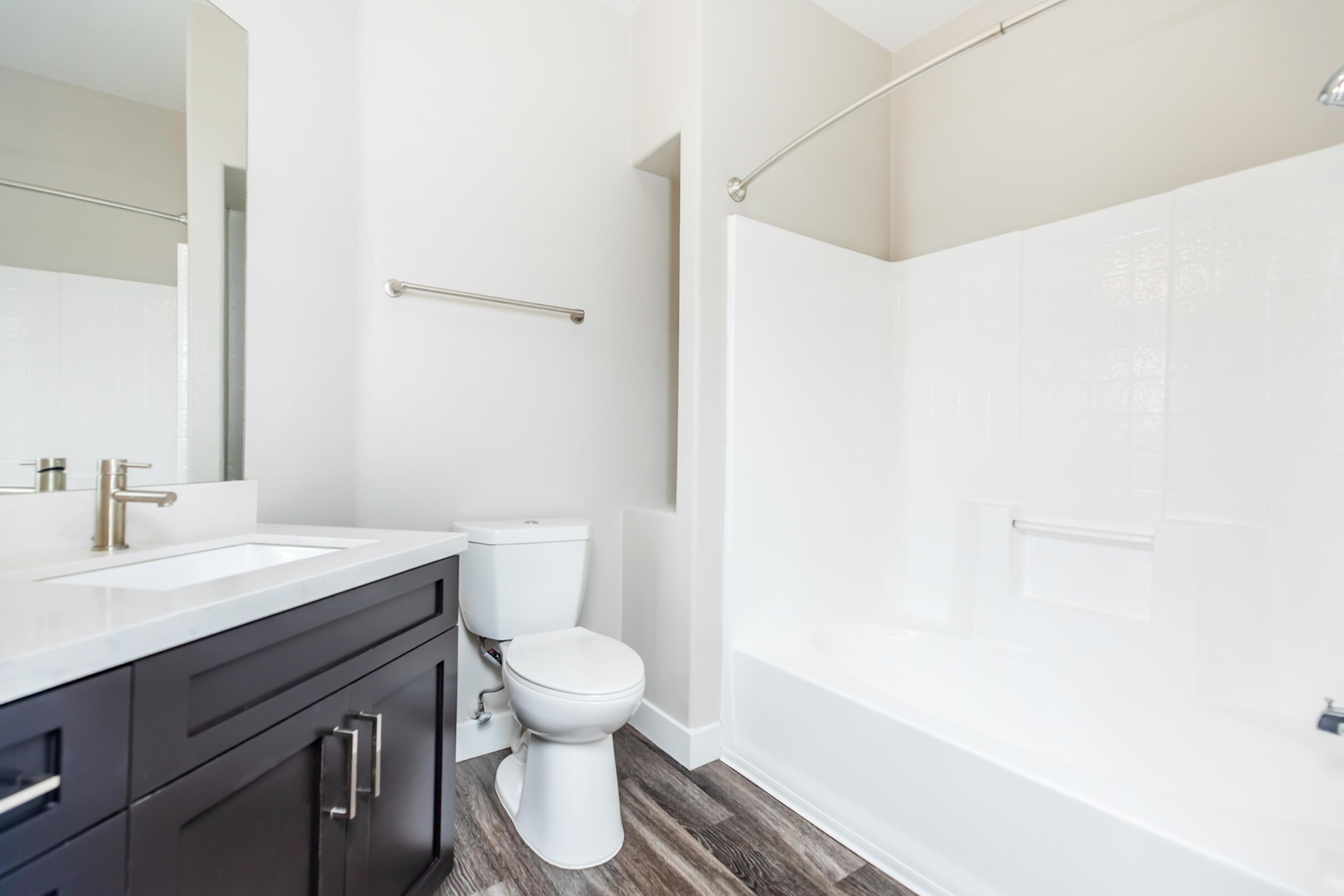 A modern bathroom featuring a white bathtub with a shower curtain rod, a wall-mounted mirror above a dark wooden vanity with a sink, and a white toilet. The floor is covered with wood-like tiles, and the walls are painted in a light neutral color.