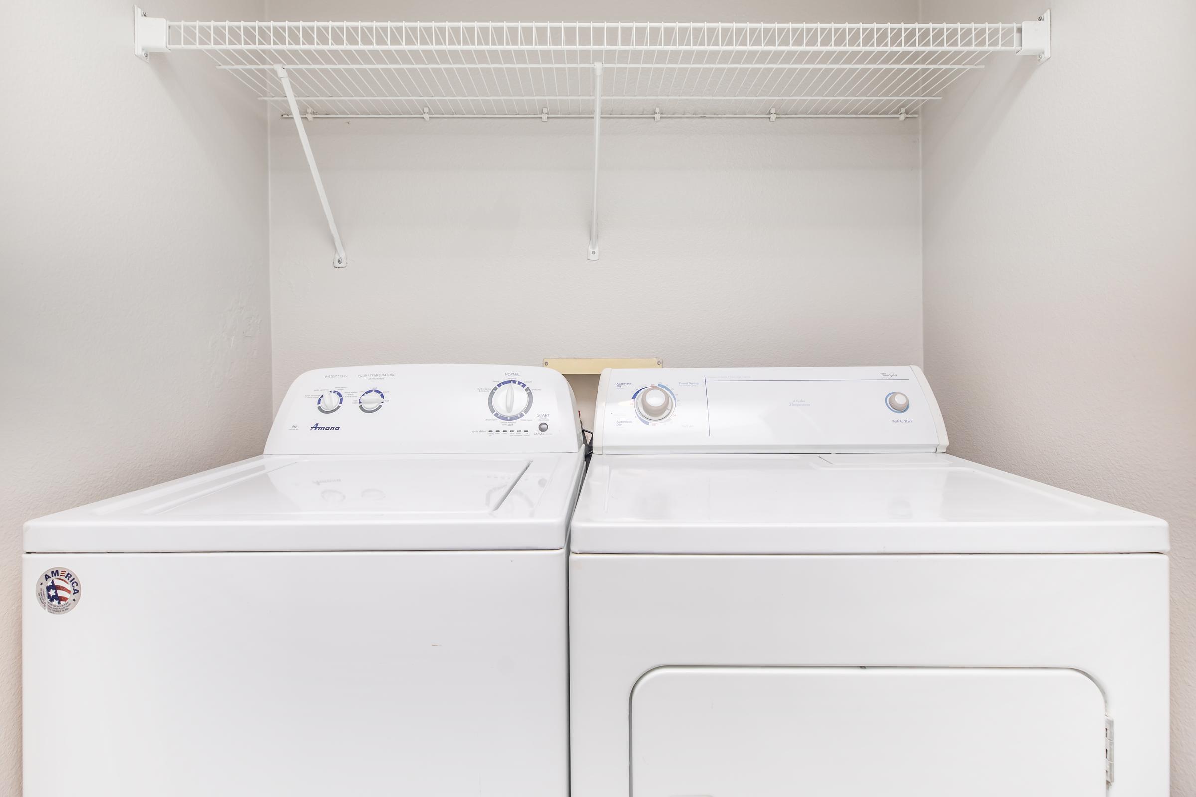 A white washing machine and dryer side by side in a laundry area, with a shelf above for storage. The machines have dials and buttons for settings, and the wall is painted a light color.