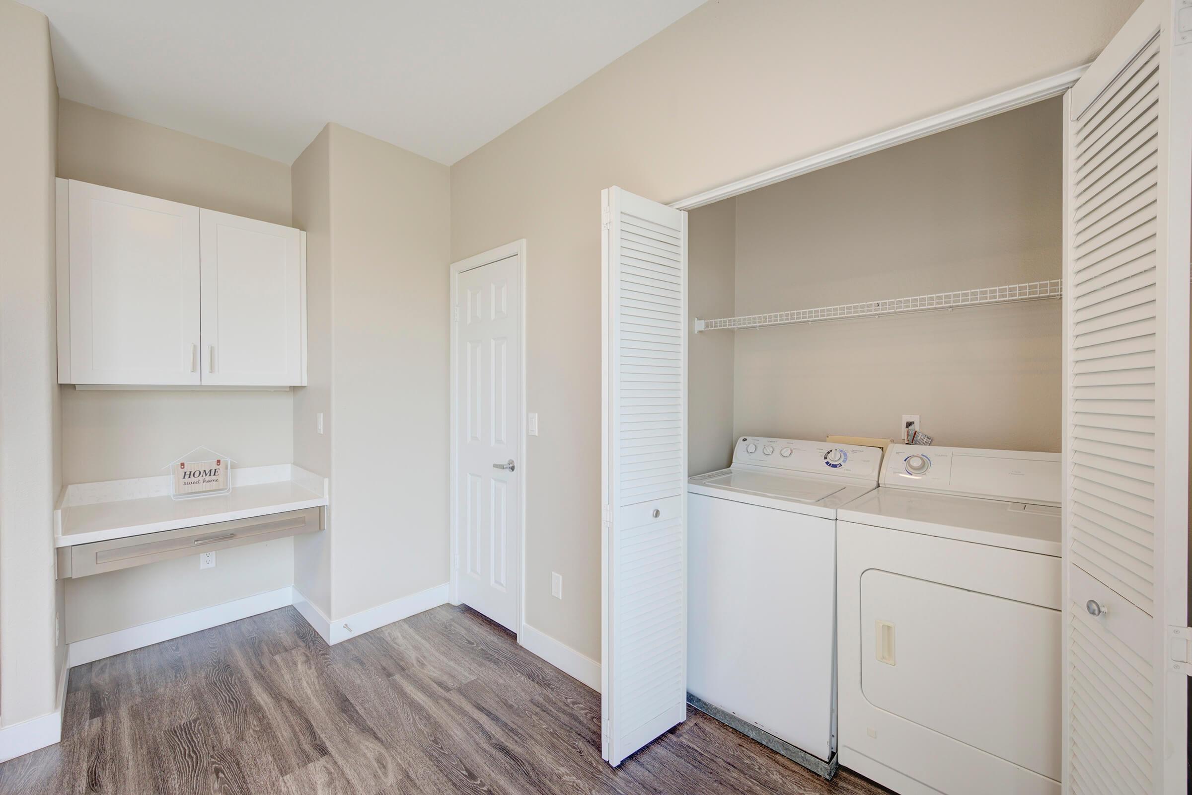 A laundry room featuring a washer and dryer in a white cabinet setup. There are light-colored walls and a built-in countertop. The flooring has a wood-like appearance, and a door leads to another room. Two white closet doors are partially open, revealing storage space.