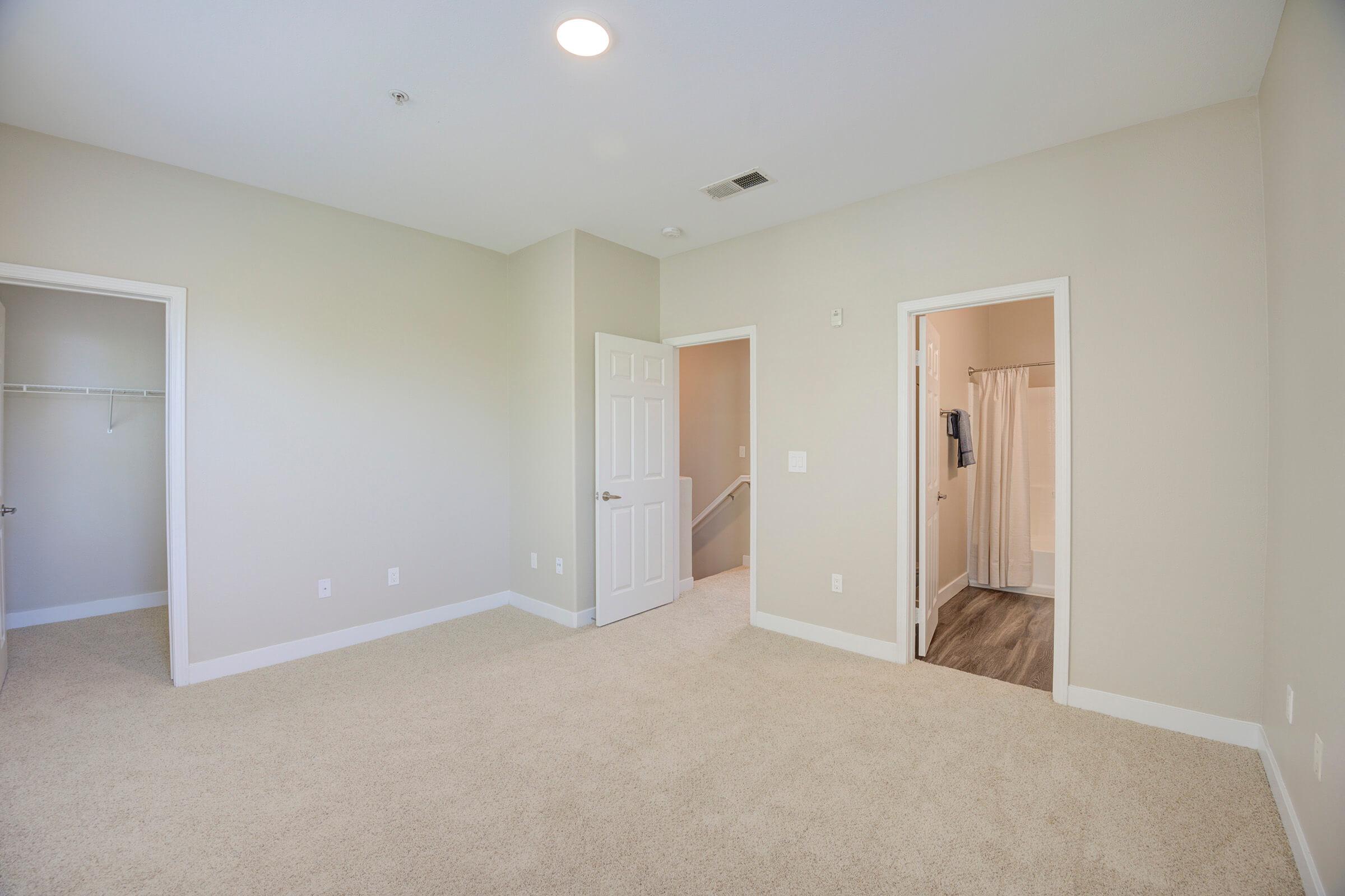 A spacious, well-lit room with beige walls and carpeted floor. There are two doorways: one leading to a closet and another to a bathroom with a shower visible through an open door. A ceiling light casts a warm glow, enhancing the open feel of the space.