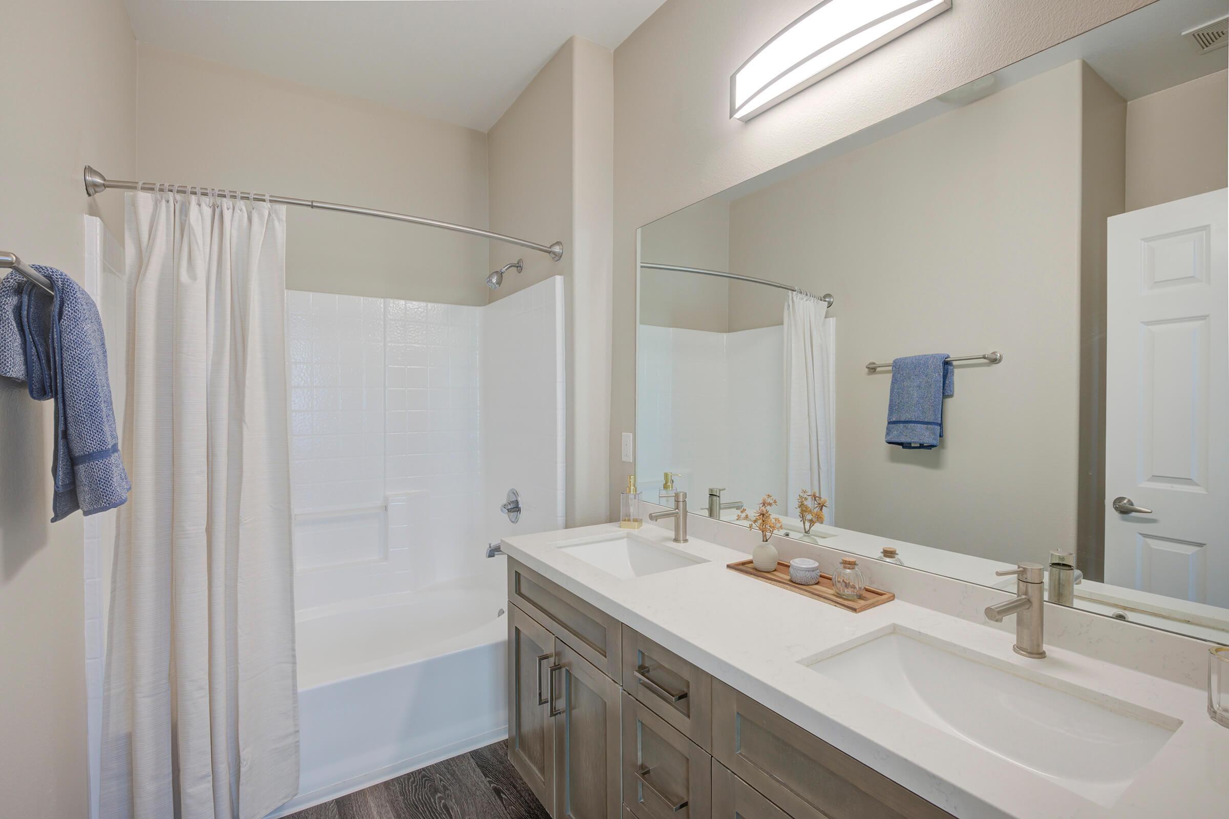 A modern bathroom featuring a double vanity with two sinks, a large mirror, and a light fixture above. The space includes a white bathtub with a shower curtain, light-colored walls, and a wooden tray on the counter with decorative items. A towel hangs on a rack, adding a touch of color to the neutral decor.