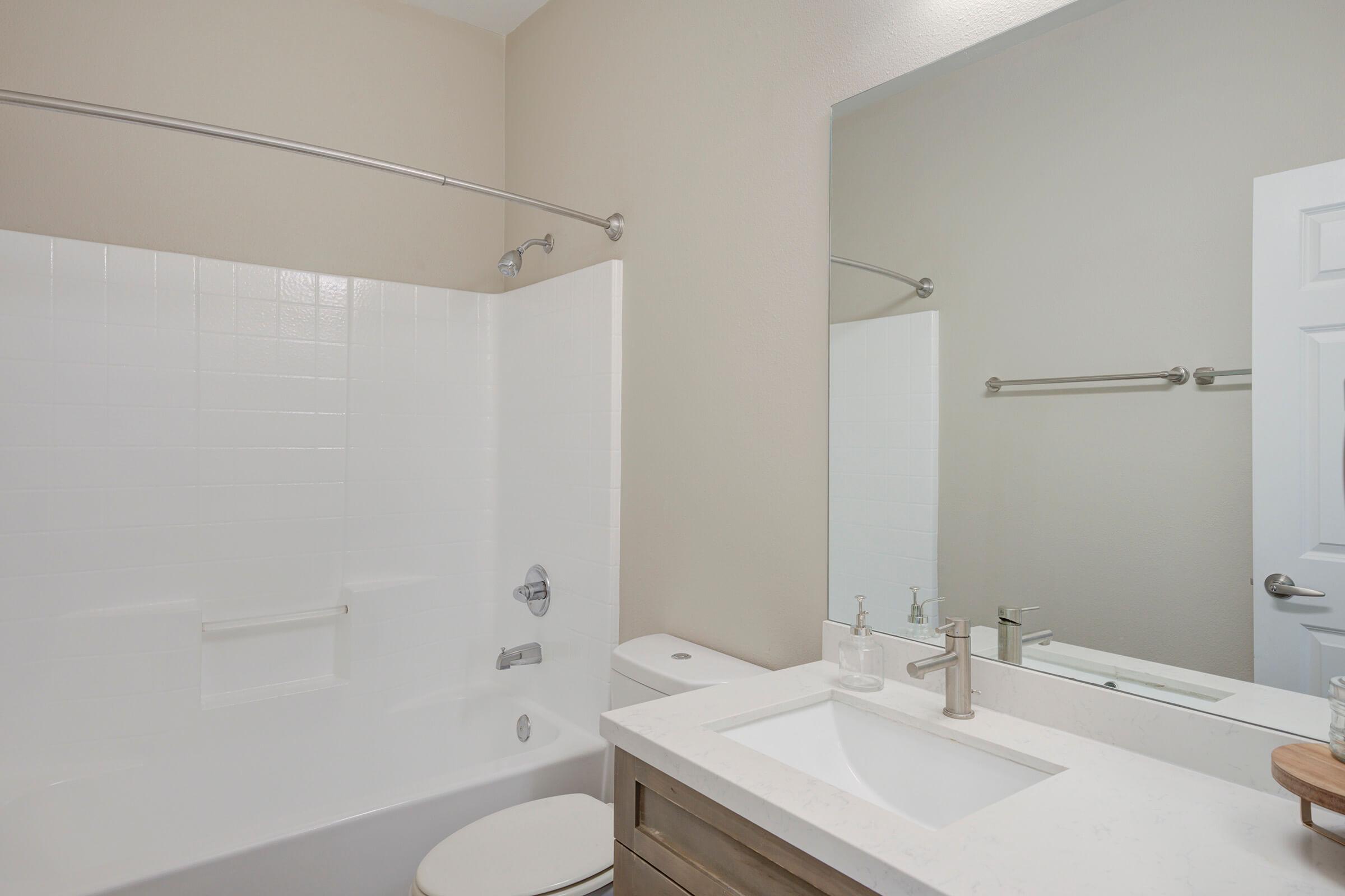 A clean and modern bathroom featuring a white bathtub with a shower curtain, a contemporary sink with a sleek faucet, and a large mirror above the sink. The walls are a light beige color, and the floor features a neutral tile. A towel bar is visible on the wall near the mirror.