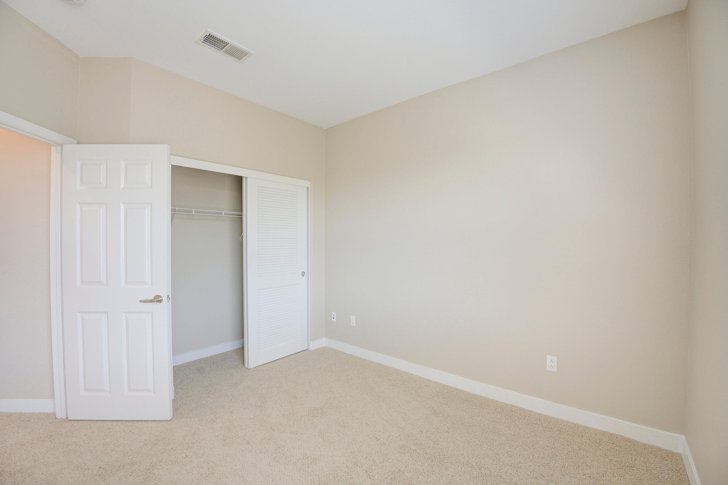 A spacious, empty bedroom featuring light beige walls and carpeting. The room includes two closet doors, one of which is a sliding door. The space is well-lit and has a minimalistic design, ideal for personalizing or furnishing.