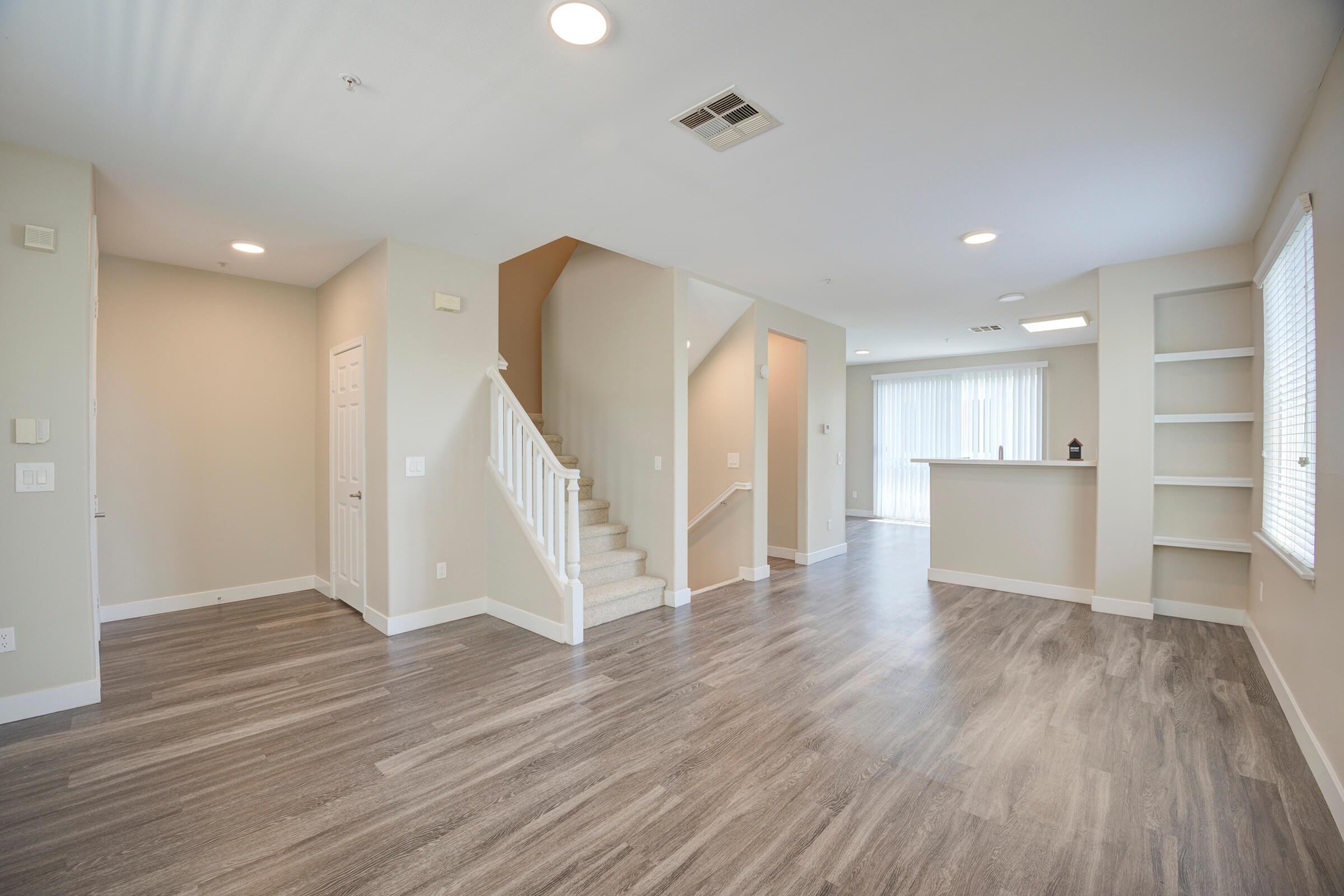 A modern, spacious interior of a home featuring light-colored walls and wood-style flooring. The image shows a living area with a staircase leading to the upper floor, an open layout towards a kitchen space, and a large window offering natural light. The design is minimalistic and inviting.