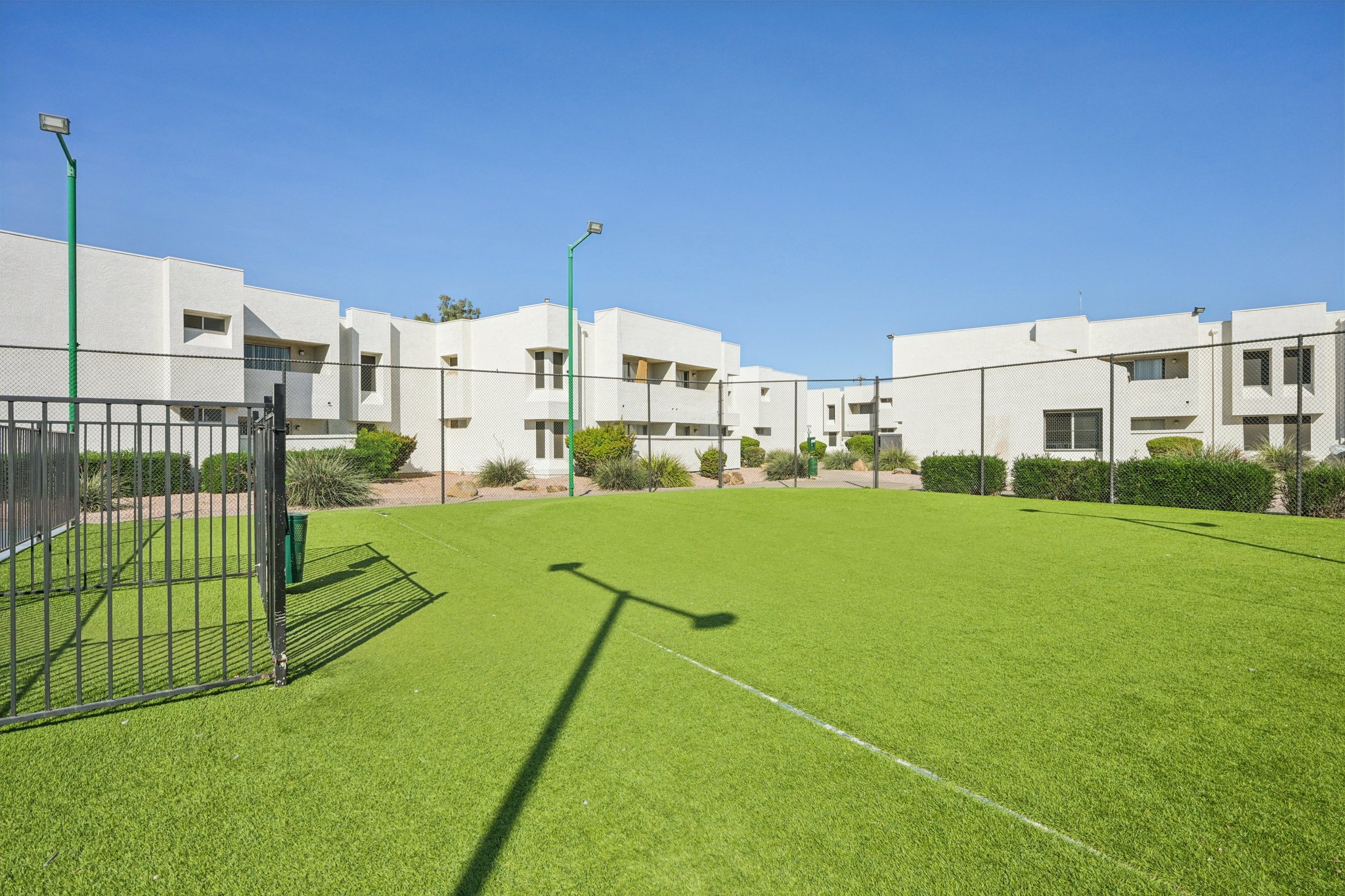 A well-maintained green lawn area with artificial turf, enclosed by a fence. Surrounding it are modern residential buildings with a clear blue sky overhead. There are several light poles and landscaped bushes along the property, creating a pleasant outdoor environment.