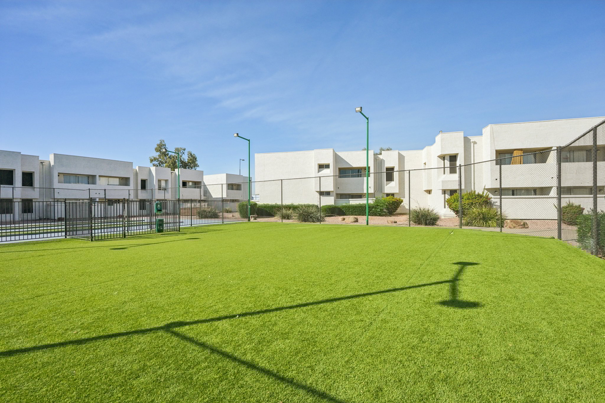 A spacious green lawn with artificial turf, bordered by a fenced area. In the background, there are white buildings with multiple windows, and tall light poles. The sky is clear and blue, indicating a sunny day.