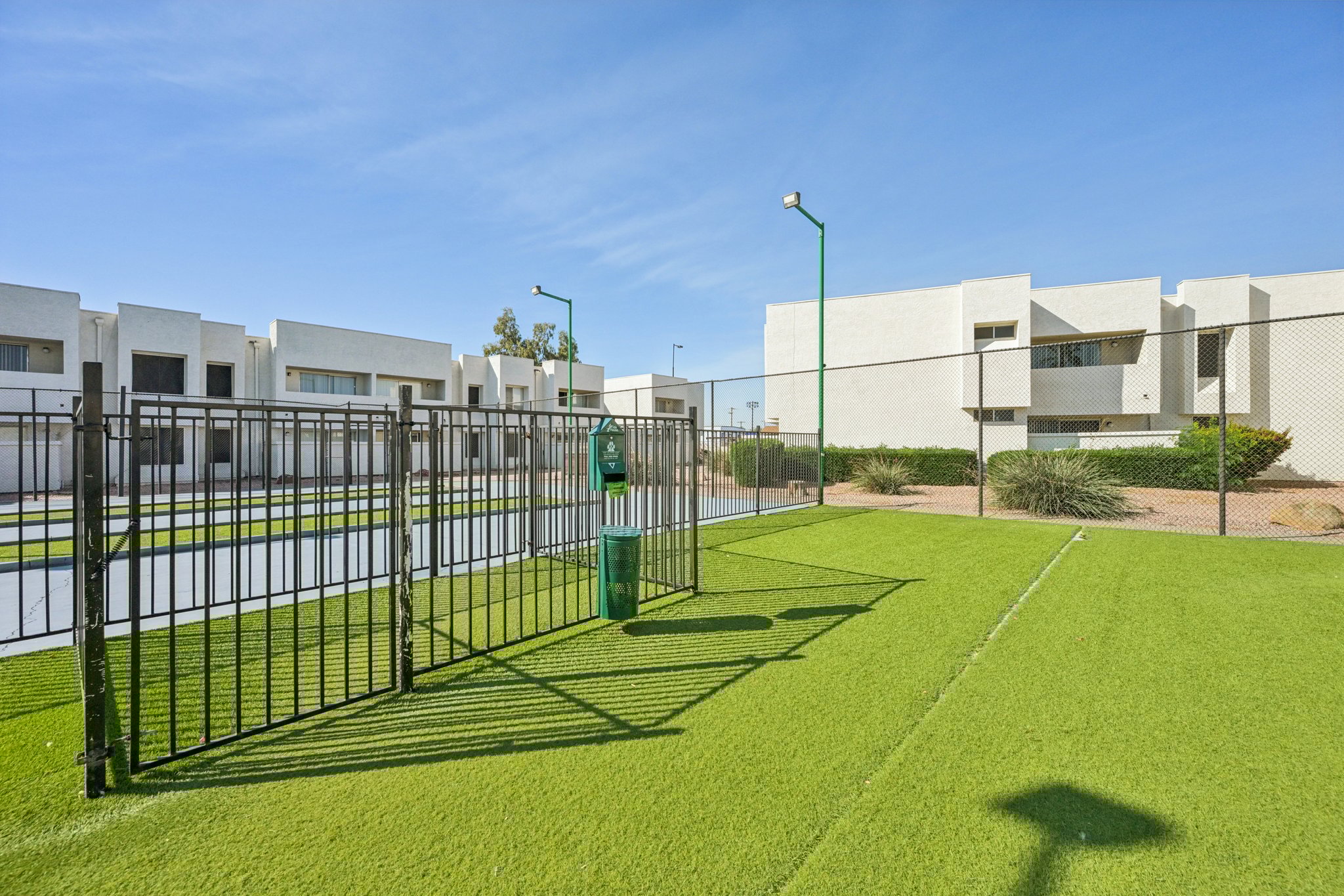 A fenced grassy area in an outdoor setting, with a green trash receptacle and nearby residential buildings. The scene features a clear blue sky and a well-maintained landscape, creating a tidy and open recreational space.
