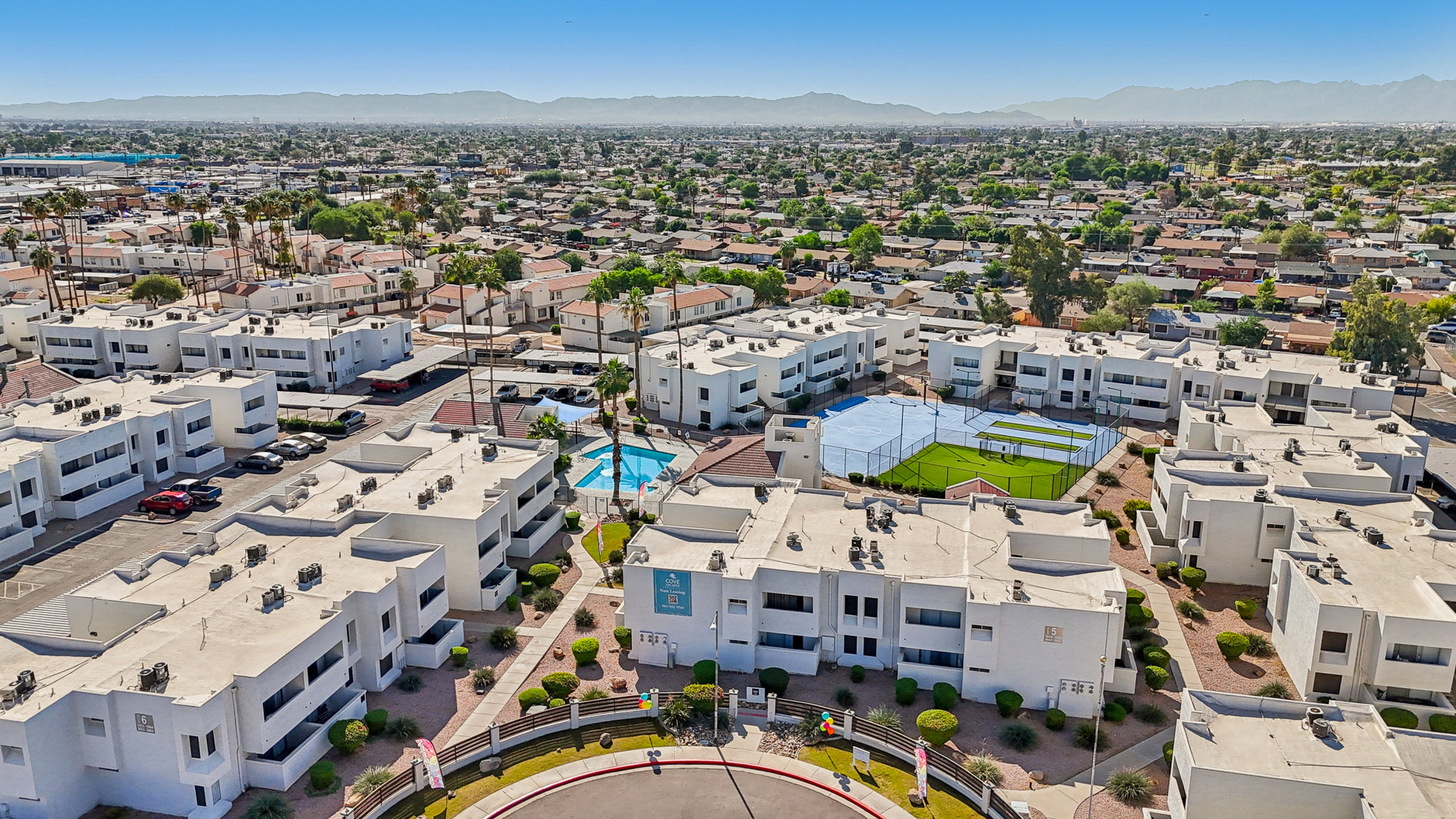 Aerial view of a neighborhood featuring white apartment buildings, a swimming pool, and well-maintained landscaping. The scene captures multiple residential units with a backdrop of distant mountains and a clear sky, showcasing a suburban environment with palm trees and a vibrant residential community.