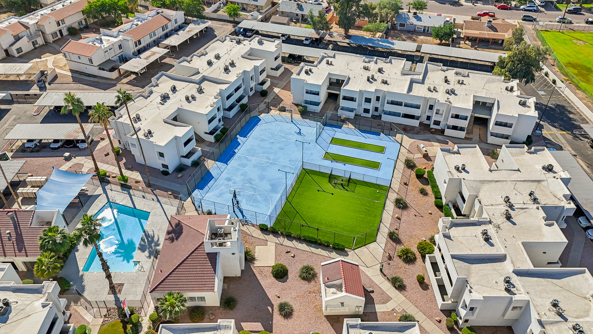 Aerial view of a residential complex featuring several white buildings with flat roofs, surrounded by landscaped areas. A blue basketball court and a green artificial turf field are visible in the center, along with a swimming pool and parking spaces. Palm trees and pathways add to the community feel.
