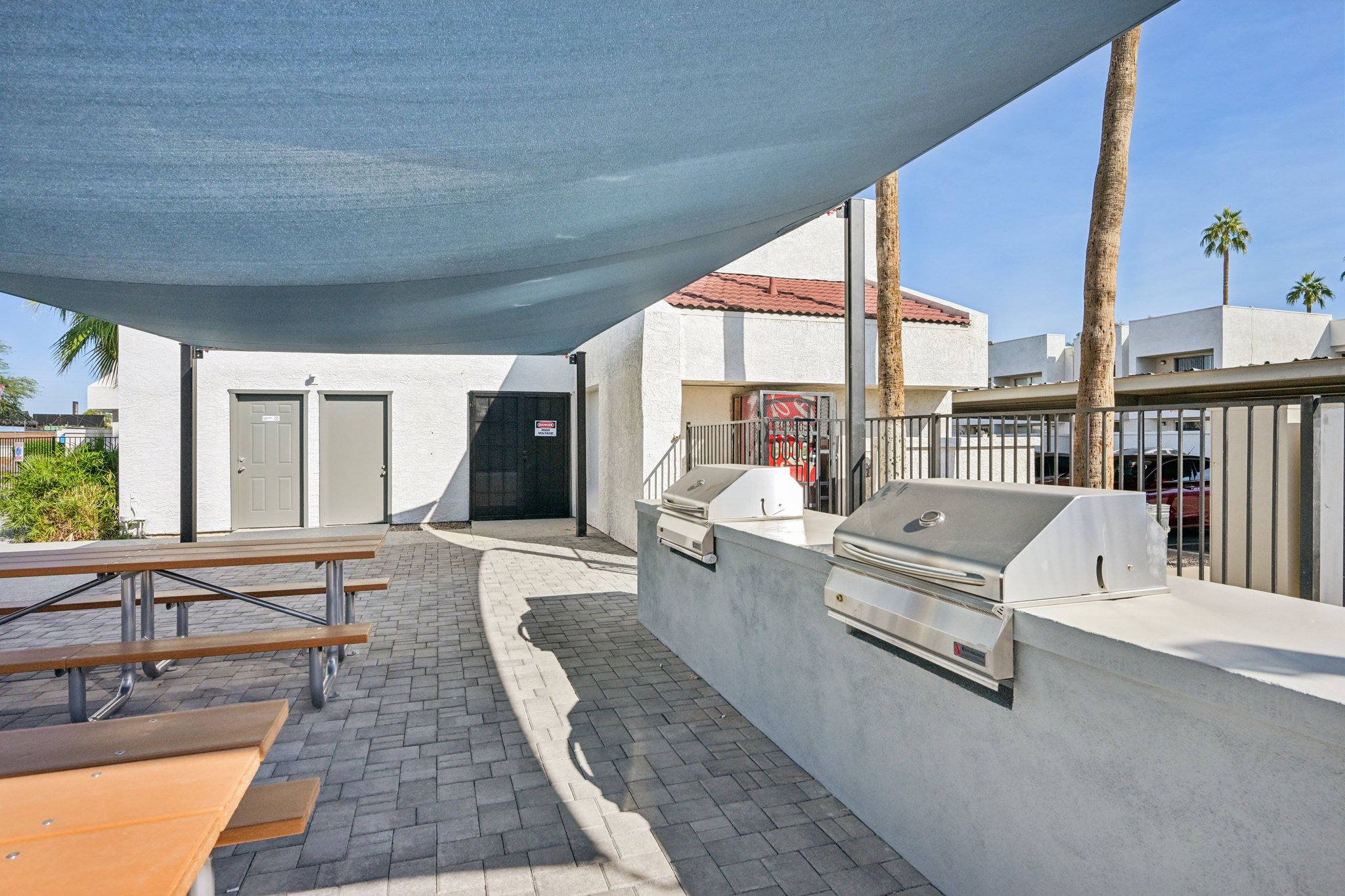 Outdoor grilling area featuring two stainless steel barbecue grills under a large shade sail, surrounded by a brick walkway and picnic tables. In the background, a building with beige walls and red roof tiles is visible, along with palm trees. The setting is designed for outdoor dining and socializing.
