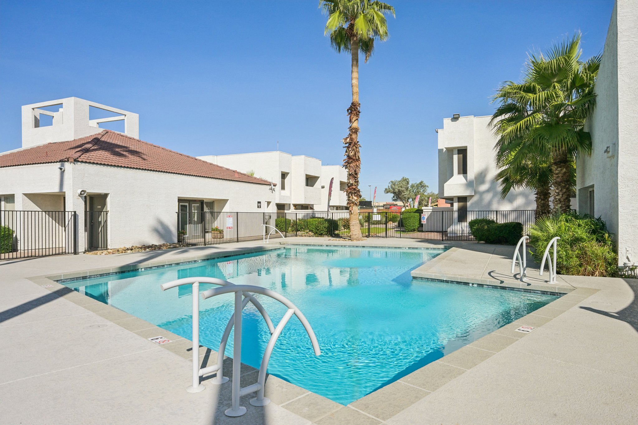 A clear swimming pool surrounded by palm trees and white buildings. The pool area features a smooth deck, stairs leading into the water, and a gated entry. Bright blue sky adds to the inviting atmosphere of the outdoor space.
