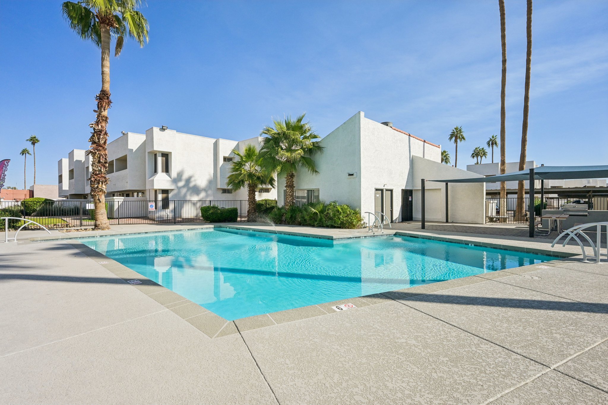 A clear blue swimming pool surrounded by palm trees and landscaping. The pool area features light-colored concrete, with lounge chairs and a shaded seating area nearby. In the background, there is a white multi-story building under a bright blue sky.