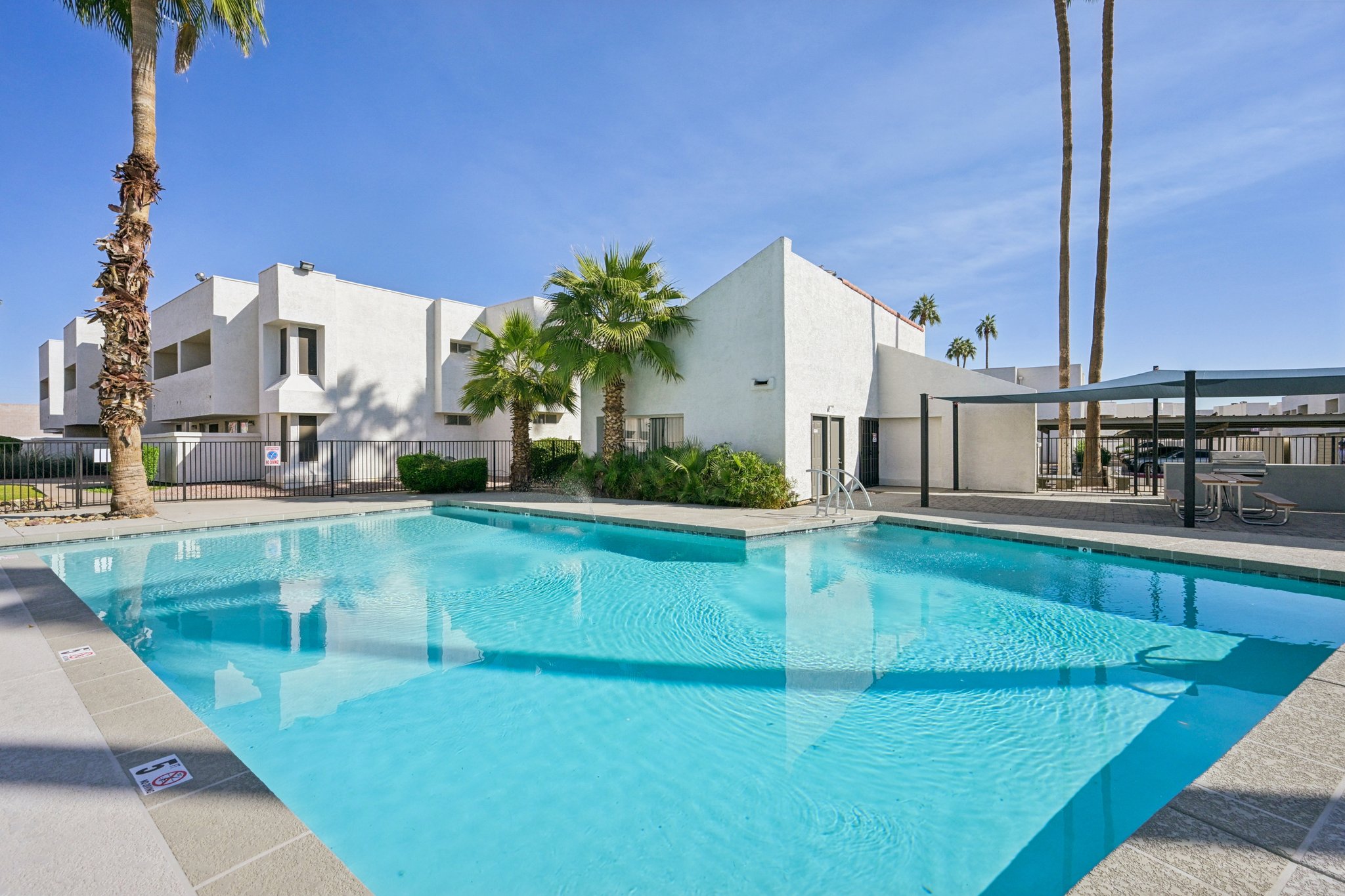 A clear blue swimming pool surrounded by palm trees and a shaded seating area. In the background, a modern white building with balconies is visible. The scene is set against a bright blue sky, indicating a sunny day.