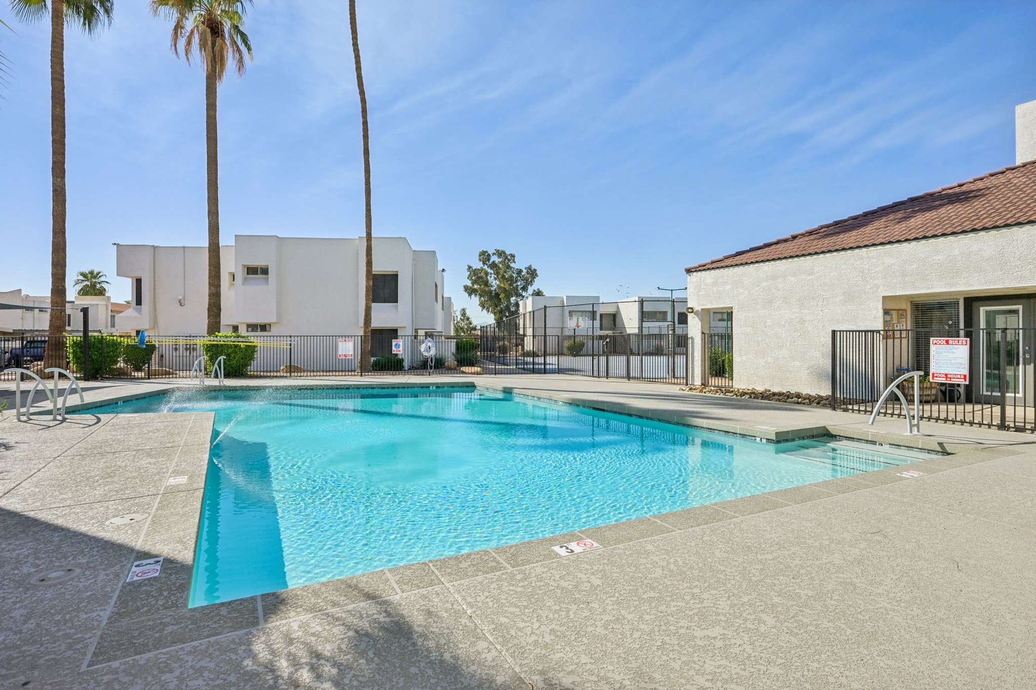 A clear blue swimming pool surrounded by a concrete deck and palm trees. The pool area features lounge chairs and a safety fence, with residential buildings in the background under a bright blue sky.
