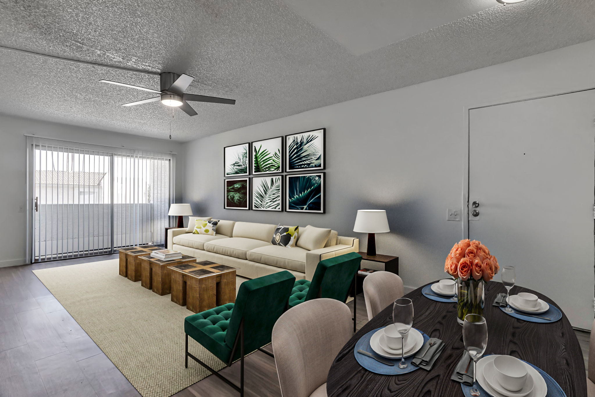 A modern living room featuring a cream sofa with decorative pillows, a large coffee table, and green accent chairs. There are four framed botanical prints on the wall, and a dining table set with plates and glasses, decorated with a vase of orange roses. A sliding door leads to a balcony, illuminated by natural light.