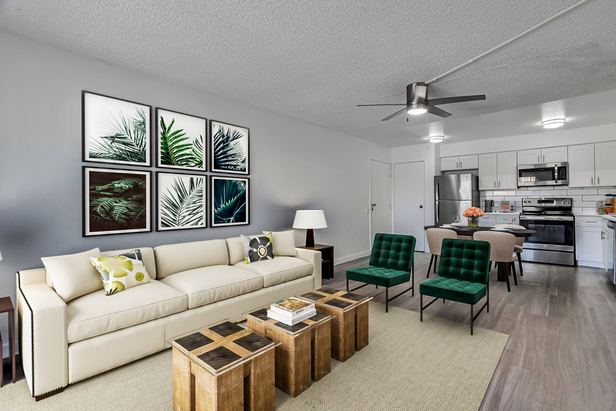A modern apartment living room featuring a beige sofa with colorful throw pillows, two green chairs, and a wooden coffee table. The walls are adorned with framed botanical prints. A dining area with a polished table and chairs is visible, alongside a kitchen area with stainless steel appliances. Bright lighting enhances the airy atmosphere.