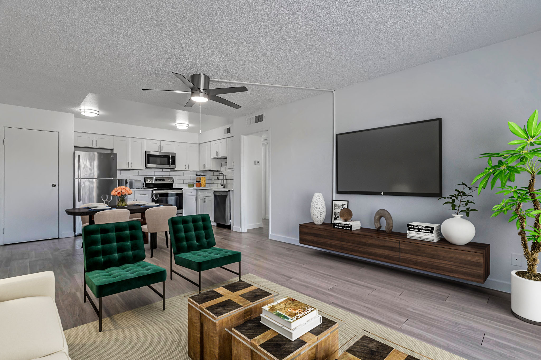 Modern living room featuring a light-colored sofa, two green chairs, and a wooden coffee table. A large wall-mounted TV is above a wooden media console with decorative items. The kitchen area is visible in the background, showcasing stainless steel appliances and white cabinetry. A potted plant adds a touch of greenery.
