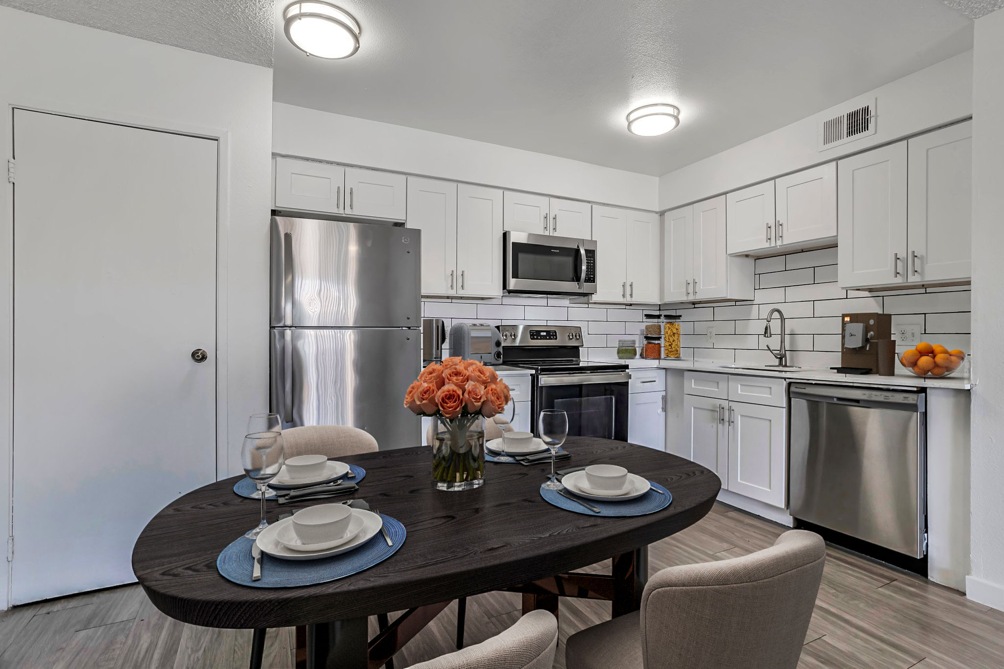A modern kitchen with stainless steel appliances, including a refrigerator, microwave, and oven. A round wooden dining table is set with plates and glasses, accompanied by a vase of orange roses. The kitchen features white cabinetry, a tiled backsplash, and an orange bowl on the counter, creating a bright and inviting atmosphere.