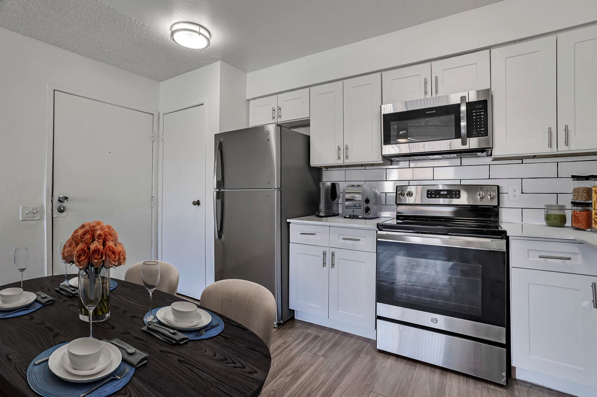 Modern kitchen with stainless steel appliances, including a refrigerator, microwave, and oven. A round dining table with a dark wood finish is set for two, featuring blue plates and a vase of orange roses as a centerpiece. The cabinets are white with a tiled backsplash, and the space is well-lit.