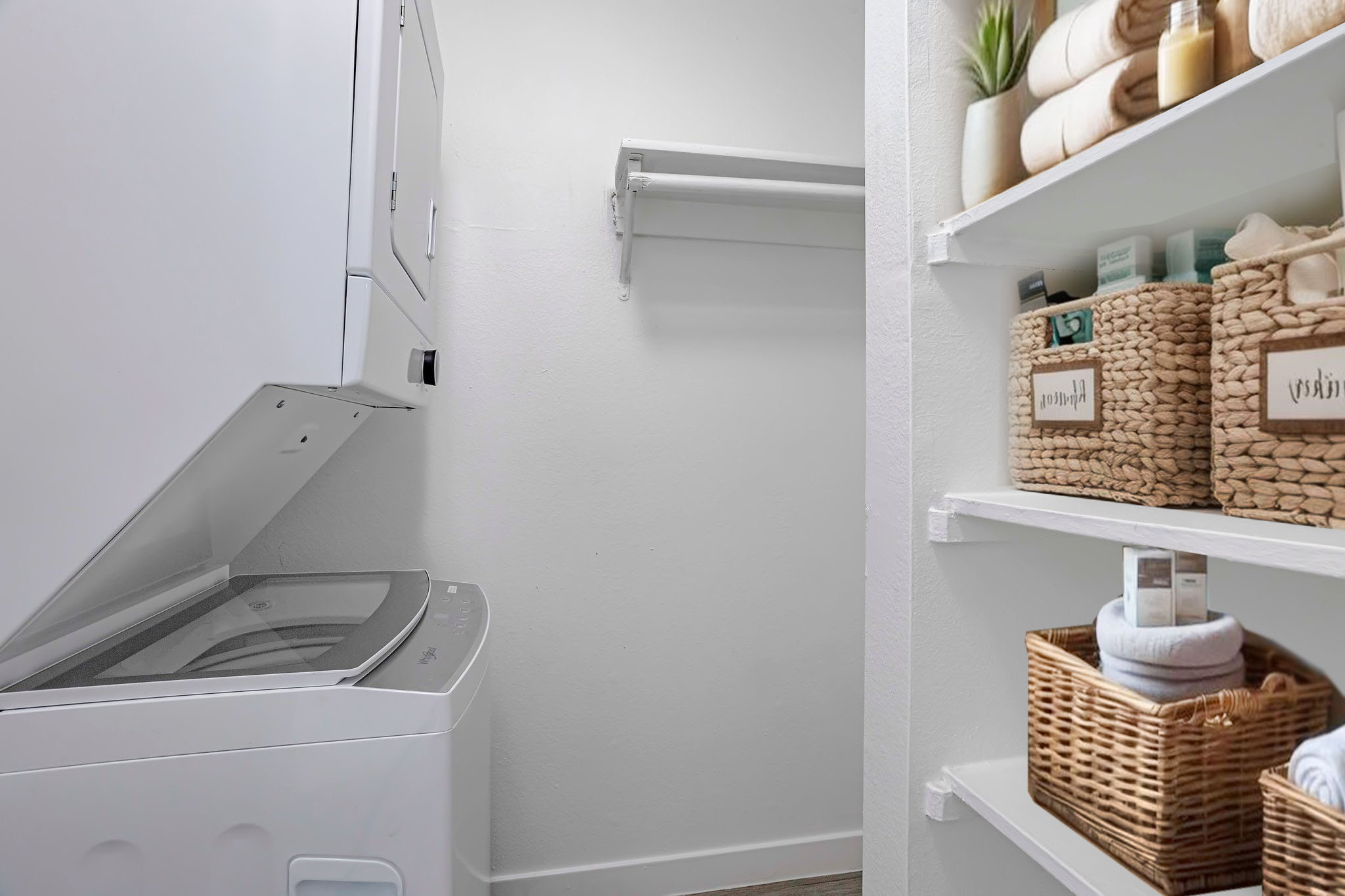 A clean laundry room featuring a stacked washer and dryer, a white wall with a shelf, and a storage area with neatly organized baskets and towels on shelves, creating a tidy and functional space.