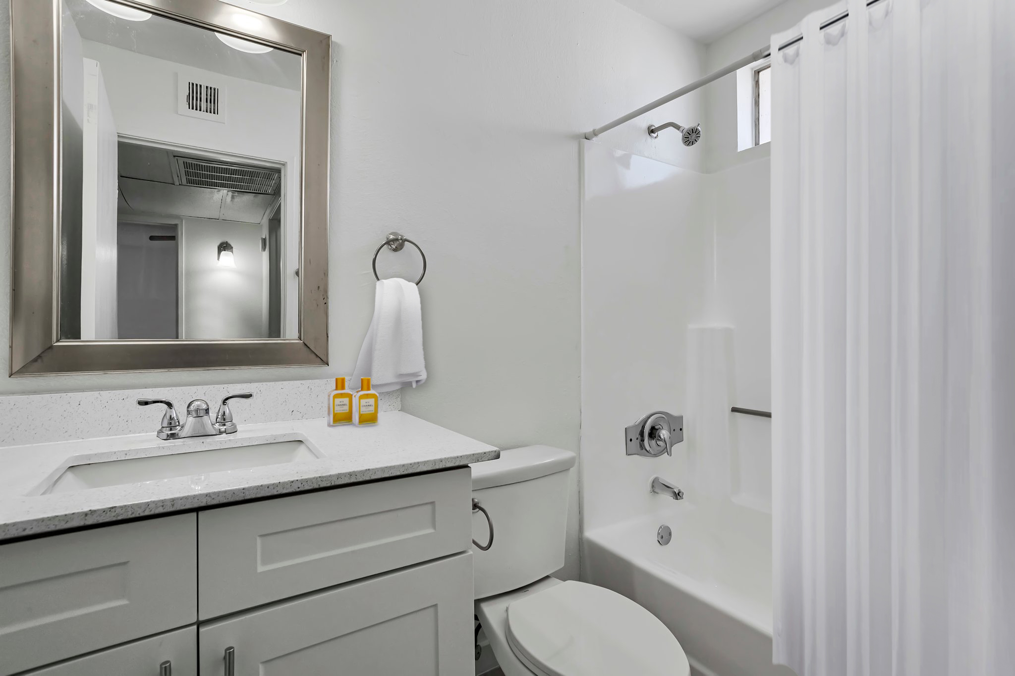 A clean and modern bathroom featuring a white bathtub with a shower curtain, a toilet, and a sink with a gray countertop. Two yellow bottles are placed near the sink, and there's a large mirror above it. Soft neutral colors enhance the bright and airy ambiance.