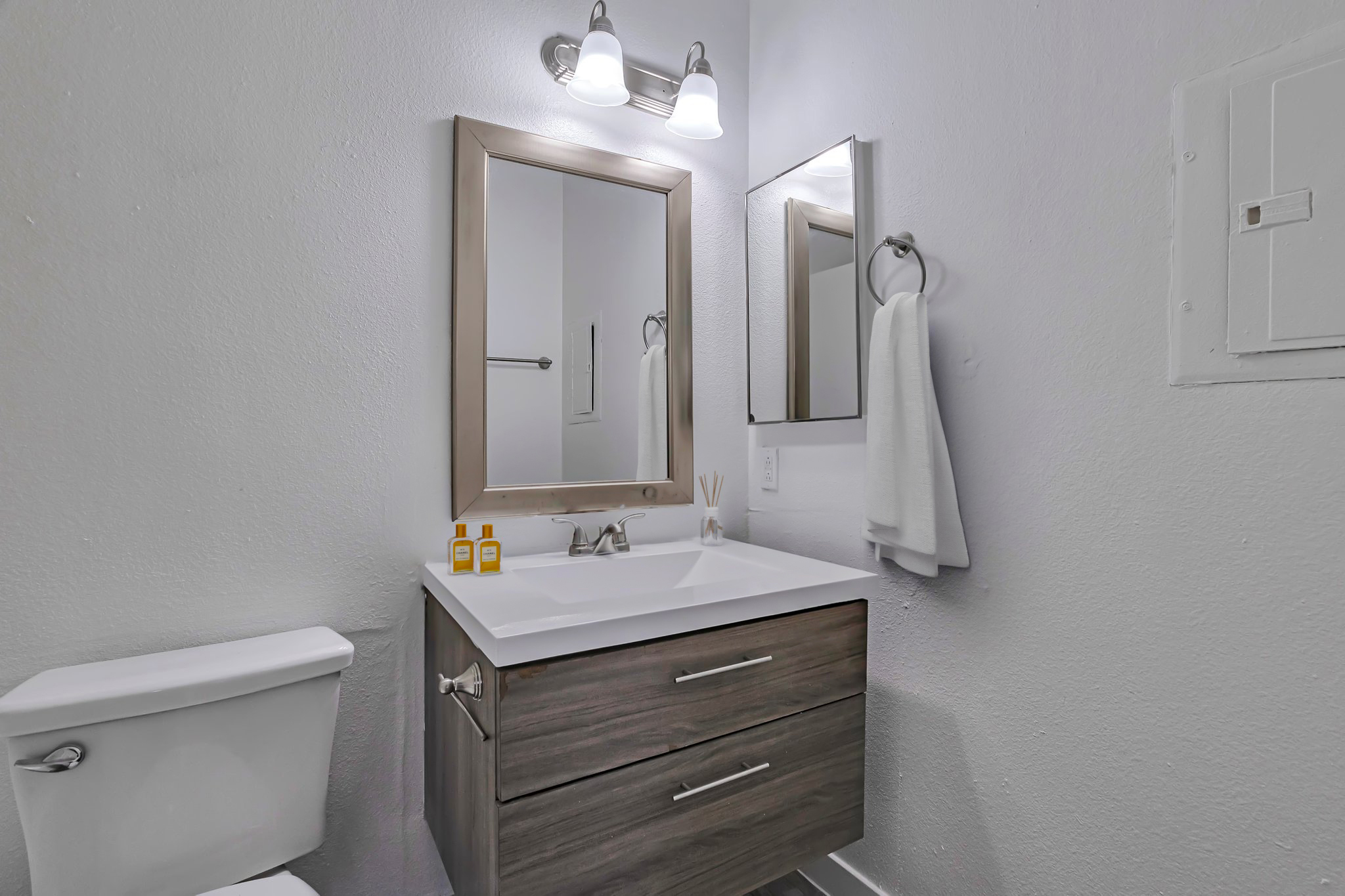 Modern bathroom featuring a white and wood vanity with a sink, two small yellow bottles on the countertop, a large mirror above, and a towel hanging on a ring. A white toilet is visible next to the vanity, with neutral-toned walls and a simple aesthetic.
