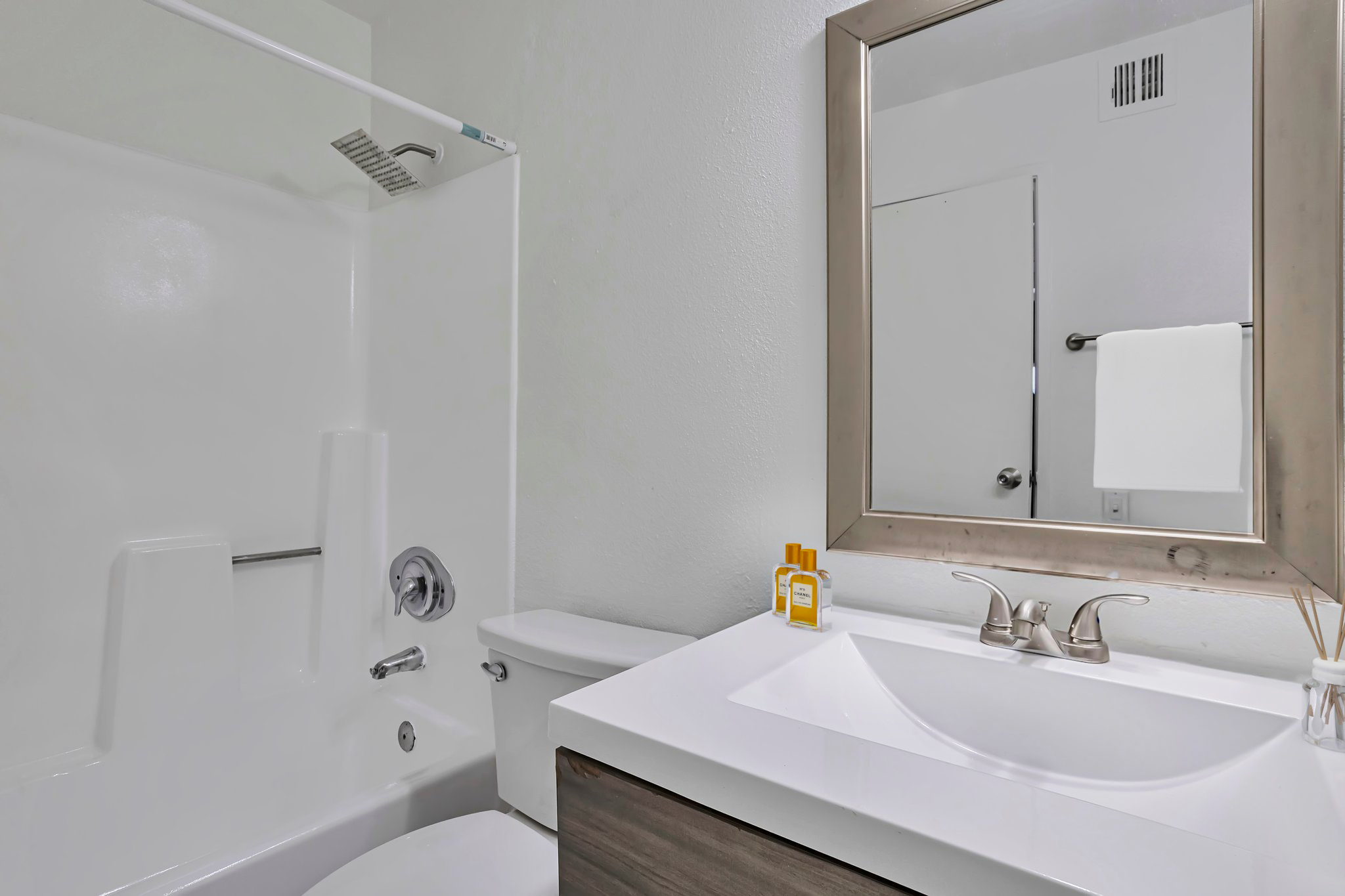 A clean and modern bathroom featuring a white bathtub and shower combo, a sleek white sink with a dark wood vanity, and a mirror with a silver frame. A towel hangs on the door, and a few toiletries are neatly arranged on the counter, creating a tidy and inviting space.