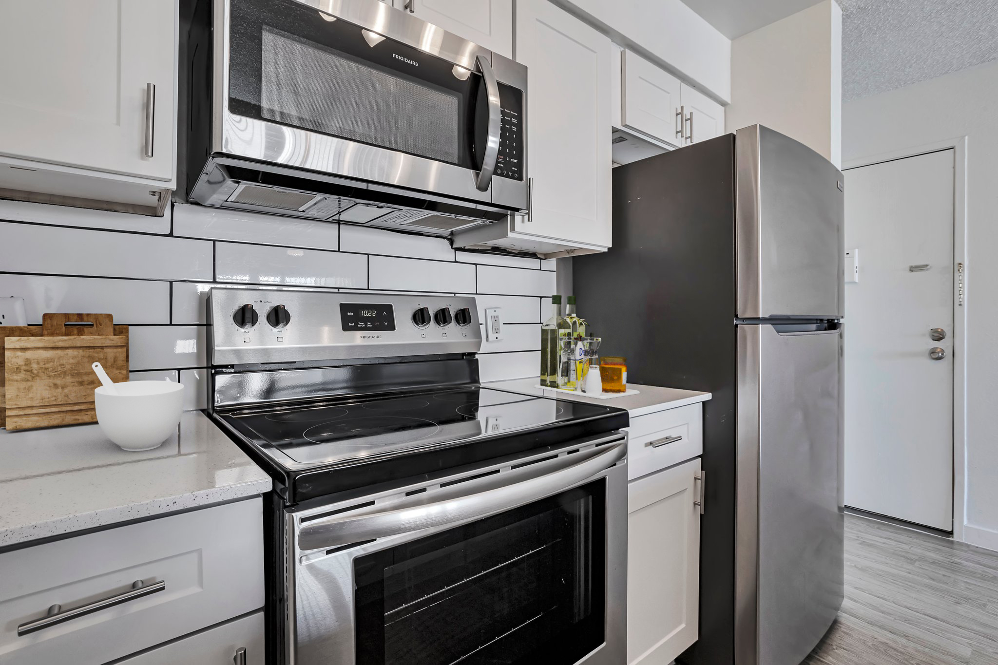 A modern kitchen featuring stainless steel appliances including a microwave, stove, and refrigerator. The cabinetry is white, with a light countertop and backsplash. A bowl and cutting board are placed on the counter, creating a clean and stylish cooking space.