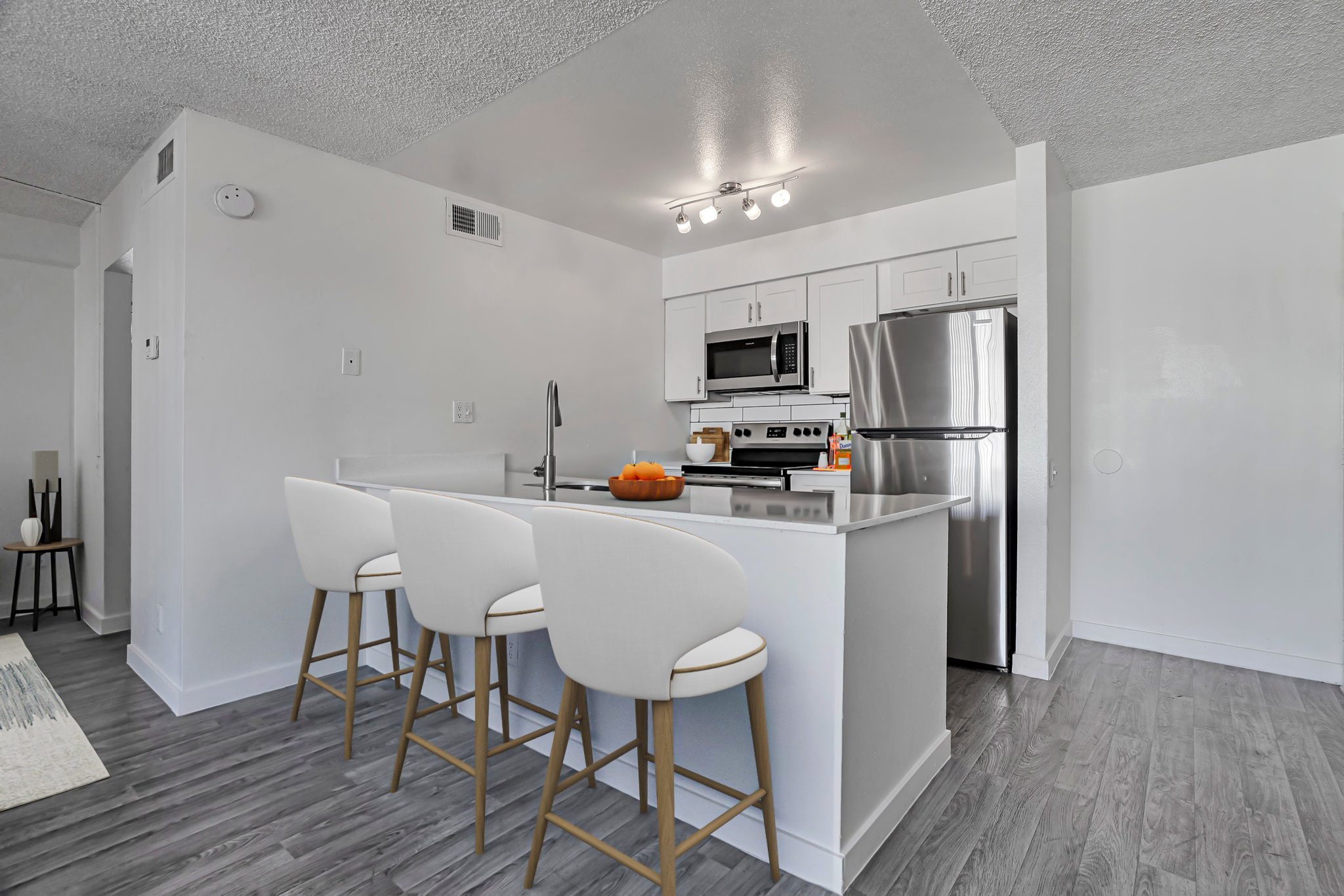 Modern kitchen featuring a spacious island with three white bar stools. The kitchen is equipped with stainless steel appliances, including a microwave and refrigerator. Bright white cabinets and countertops create a clean, contemporary aesthetic, illuminated by overhead lights. Hardwood-style flooring completes the look.