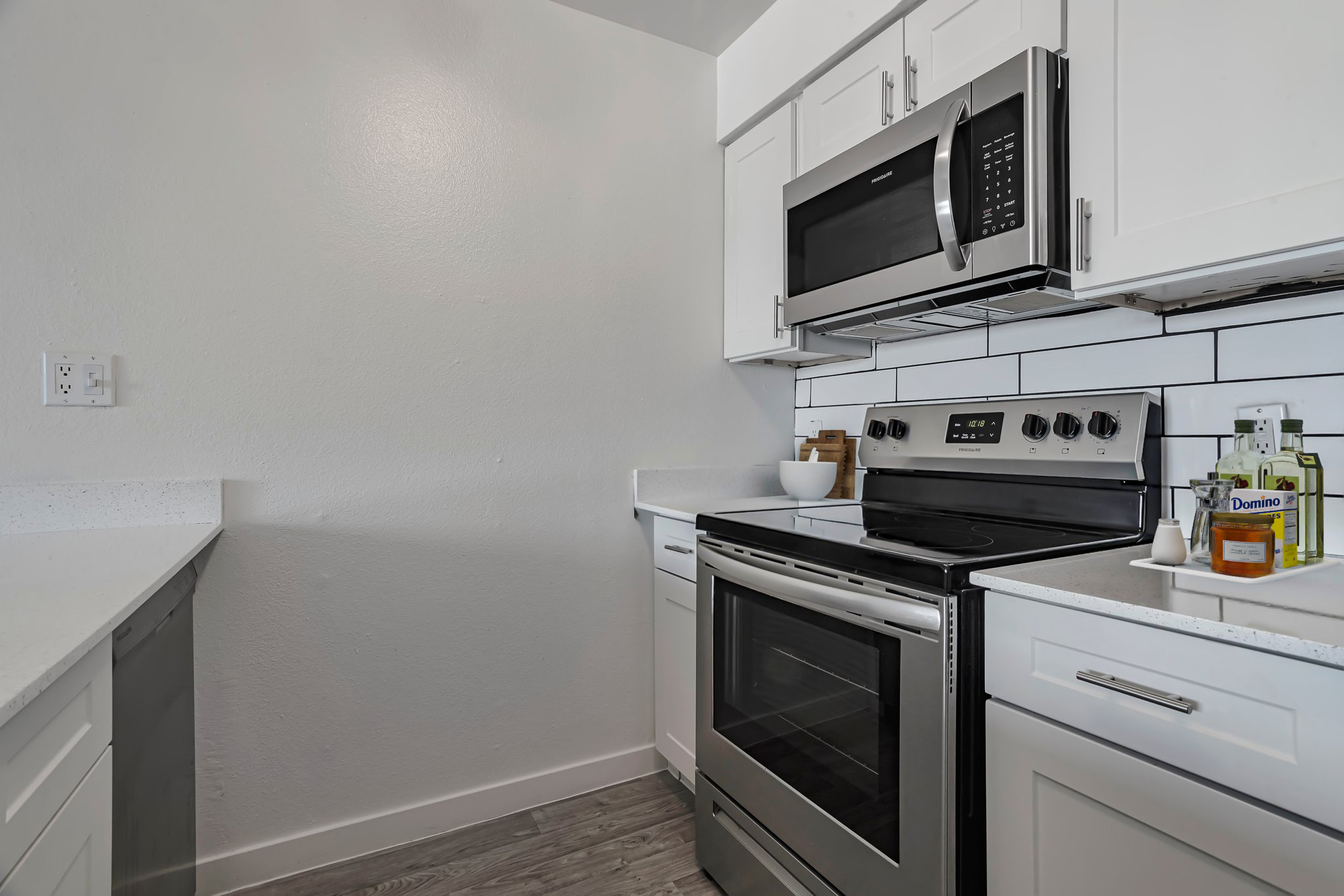 Modern kitchen featuring white cabinetry, a sleek black oven and stovetop, a stainless steel microwave above, and a white countertop. The backsplash is tiled in white, adding a clean look. There are kitchen essentials like cooking oil and seasonings on the counter, contributing to a functional space.