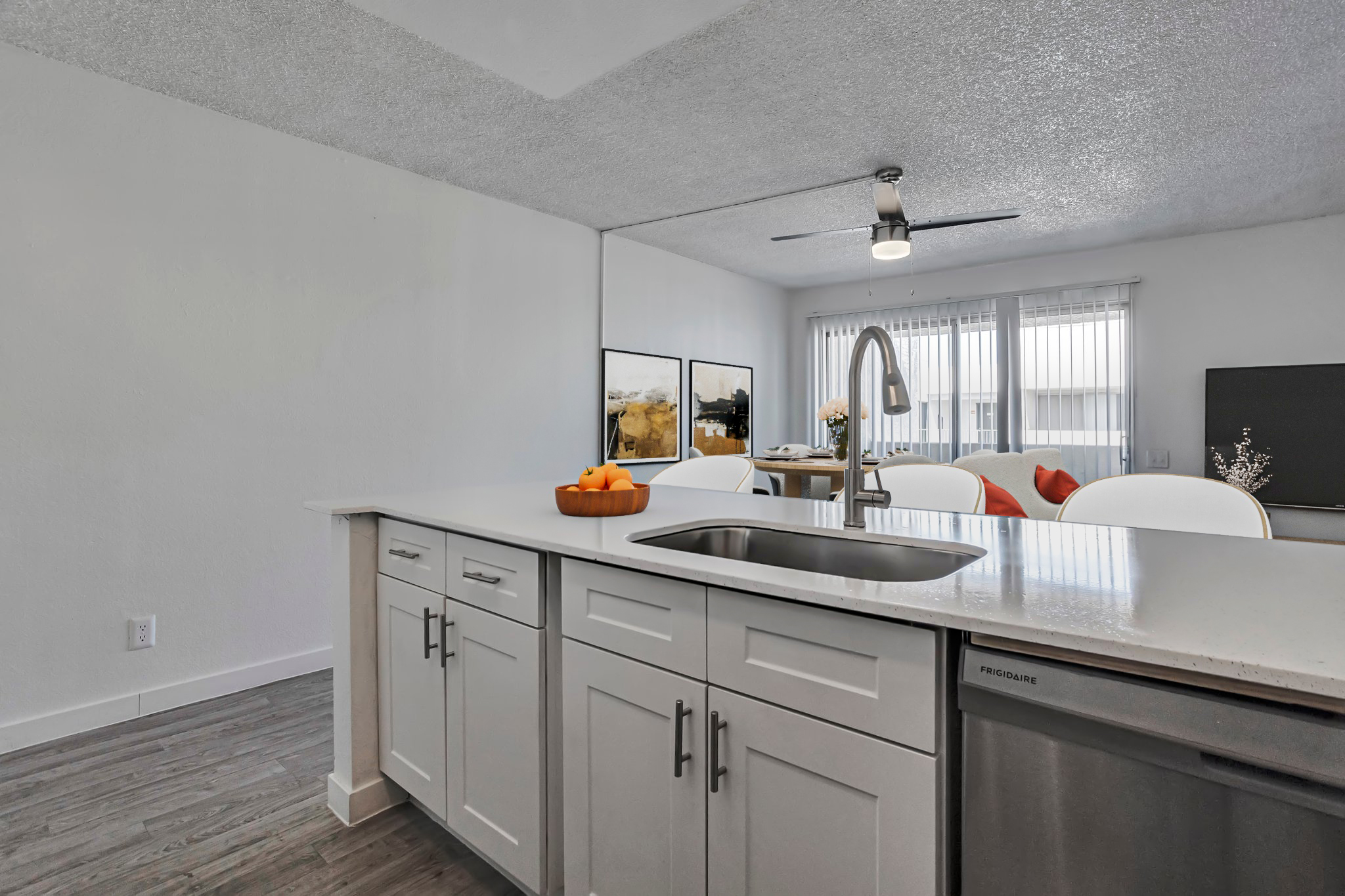 Modern kitchen with white cabinetry and a stainless steel sink. In the foreground, a bowl of oranges sits on the countertop. The background features a dining area with light-colored chairs and a living space with a television and decorative elements. The room is well-lit with a ceiling fan and large windows.