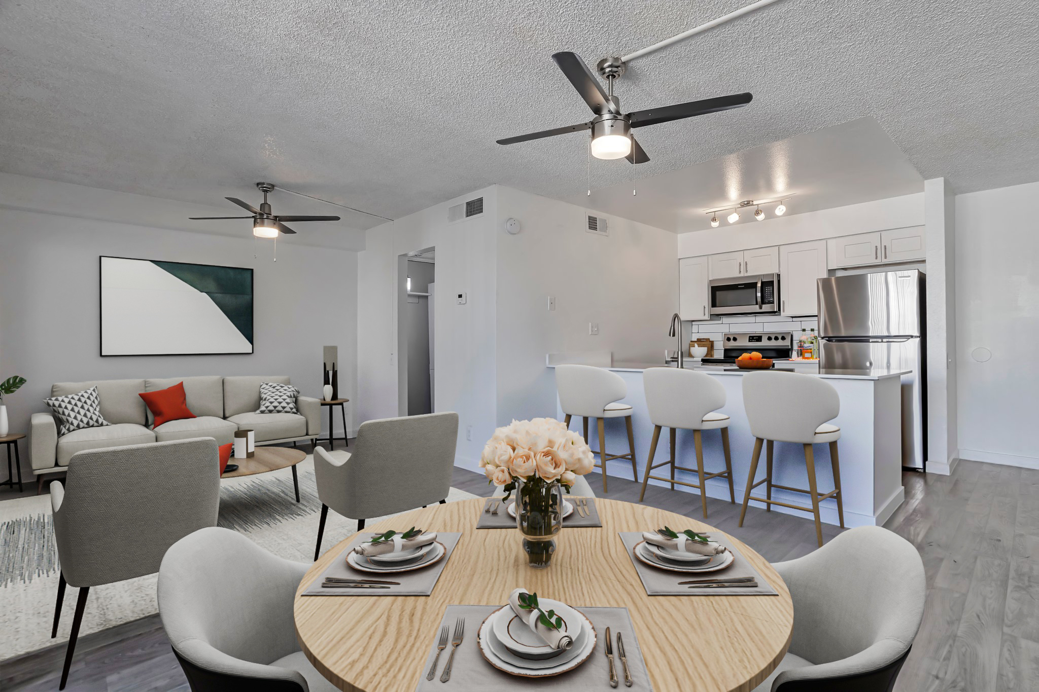 A modern, stylish apartment interior featuring a dining table set for four with white dishware and a floral centerpiece. In the background, a cozy living area with a gray sofa, two ceiling fans, and a kitchen with stainless steel appliances and a breakfast bar. Large windows allow natural light to brighten the space.
