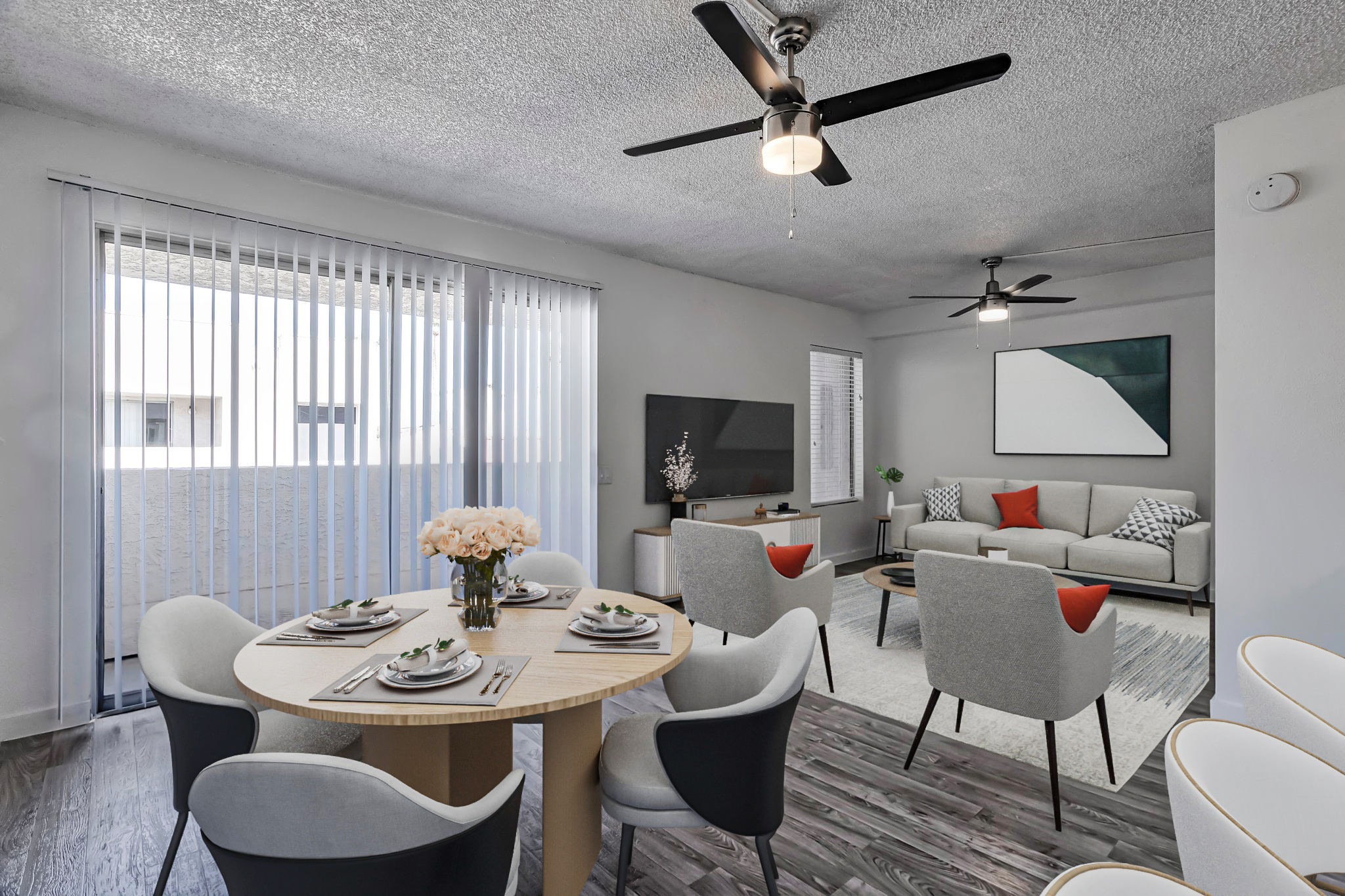 A modern living and dining area featuring a round wooden table with set dining ware, stylish chairs, and a cozy sofa adorned with colorful cushions. Large windows with vertical blinds allow natural light, complemented by ceiling fans. The space has a contemporary aesthetic with neutral tones and minimal decor.